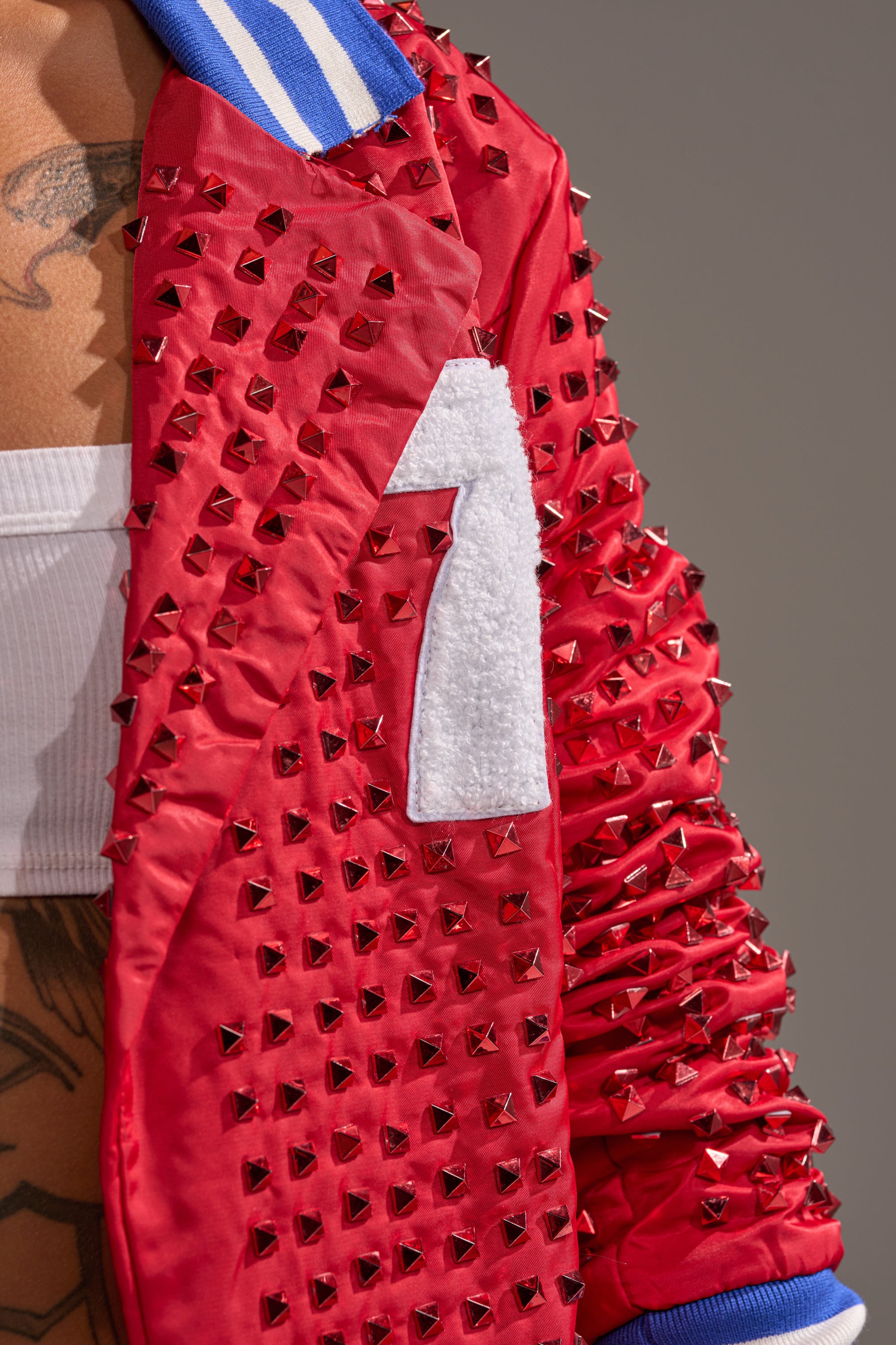 A close-up of the FIRST ROUND DRAFT PICK STUDDED BLAZER IN RED, featuring metallic studs, a white number 7 patch, and a blue-and-white striped collar. A tattooed torso in a white top is partially visible.