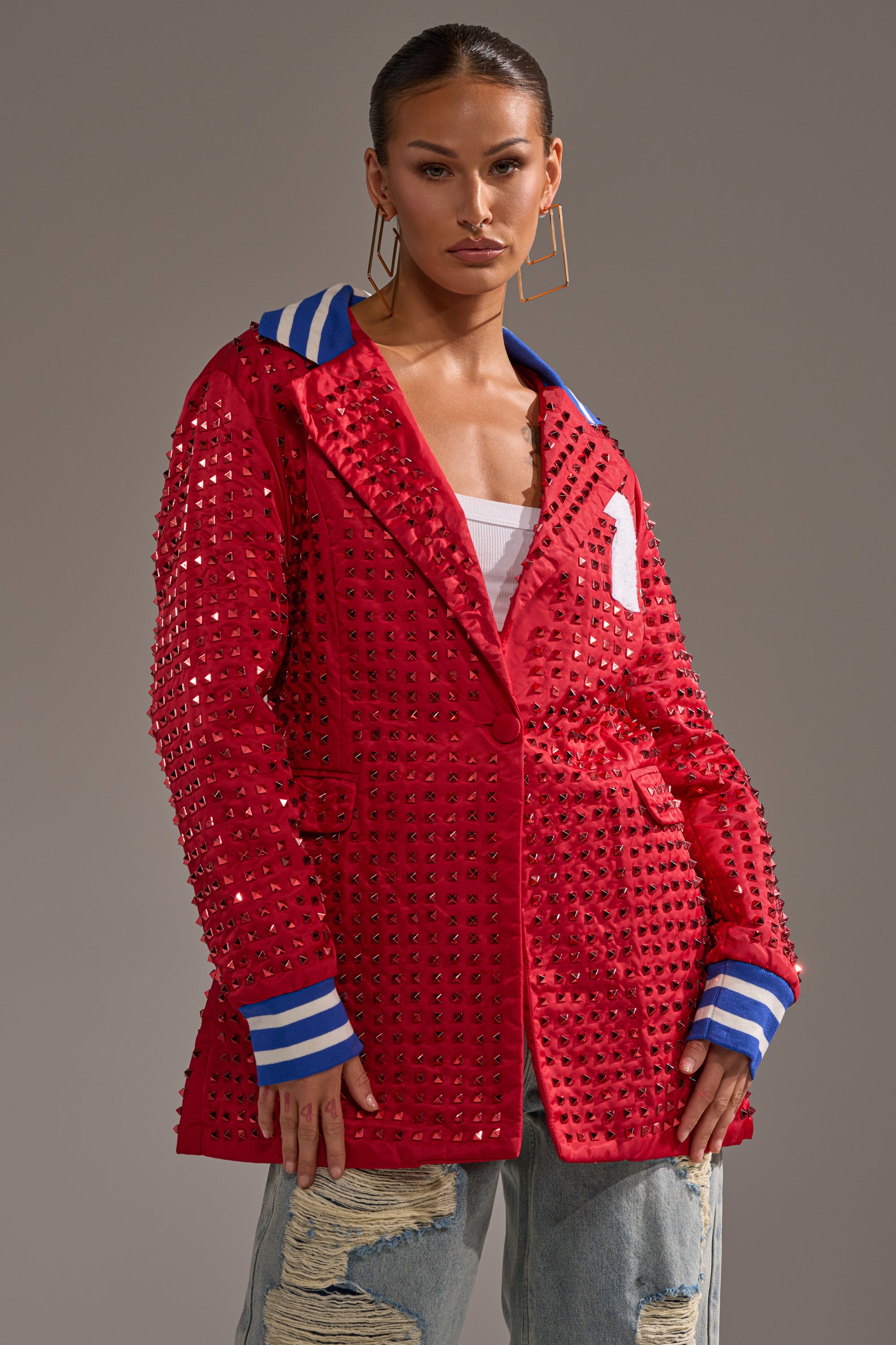 A woman confidently wears the FIRST ROUND DRAFT PICK STUDDED BLAZER IN RED, featuring blue and white striped cuffs and collar, paired with a white top, distressed light-wash jeans, and large square earrings.