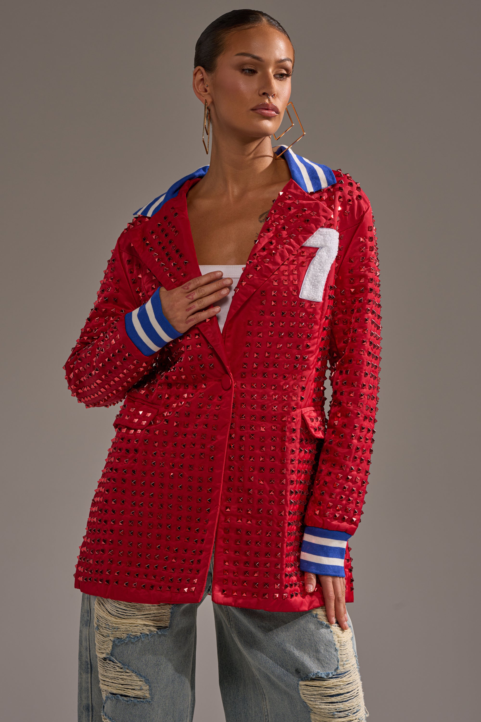 A person models the FIRST ROUND DRAFT PICK STUDDED BLAZER IN RED, featuring a blue and white striped collar and cuffs, a bold "1" on the chest, paired with hoop earrings and ripped light blue jeans against a plain background.