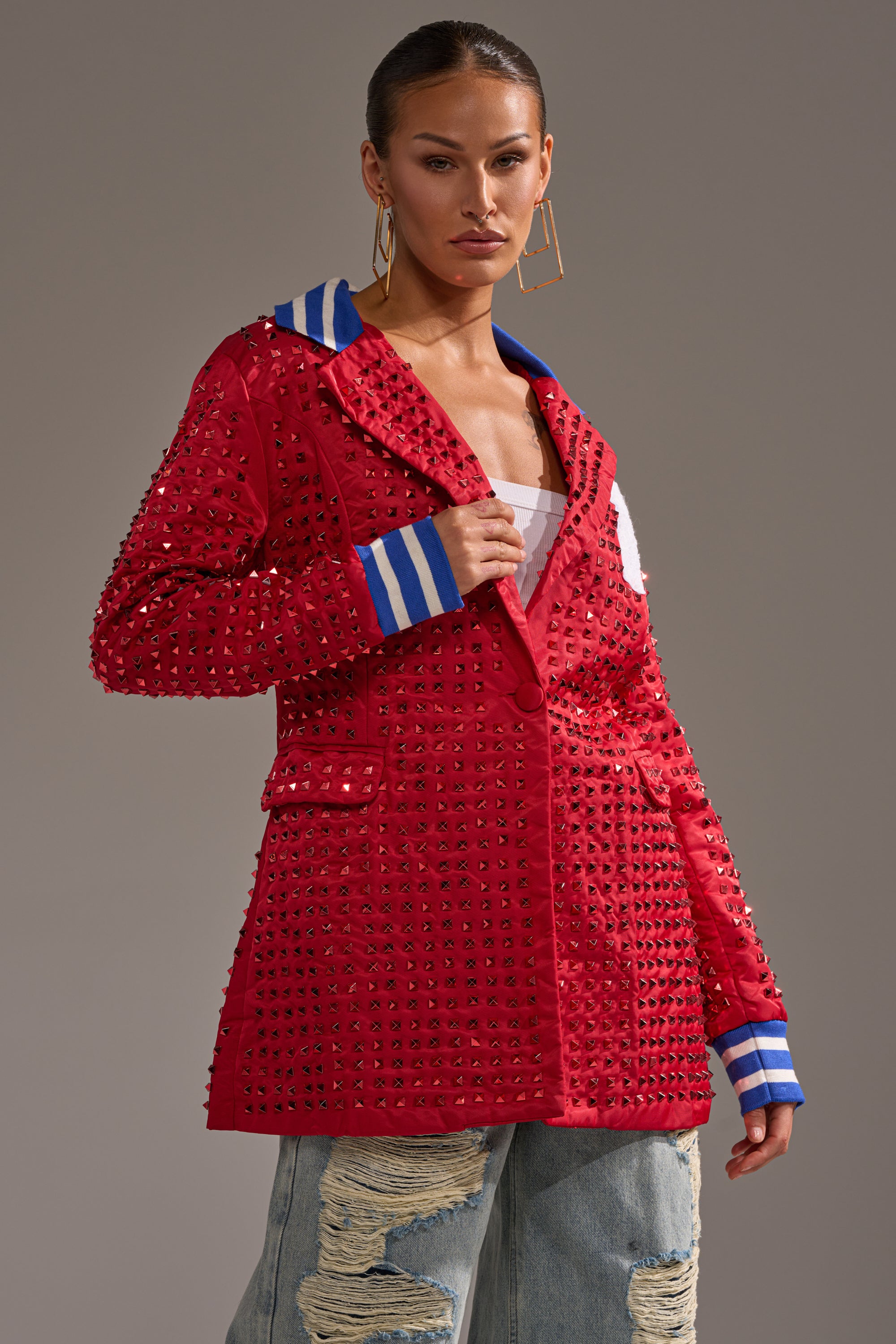 A woman poses confidently in the FIRST ROUND DRAFT PICK STUDDED BLAZER IN RED, featuring blue and white striped cuffs and collar, styled with a white top and ripped light blue jeans against a plain backdrop.
