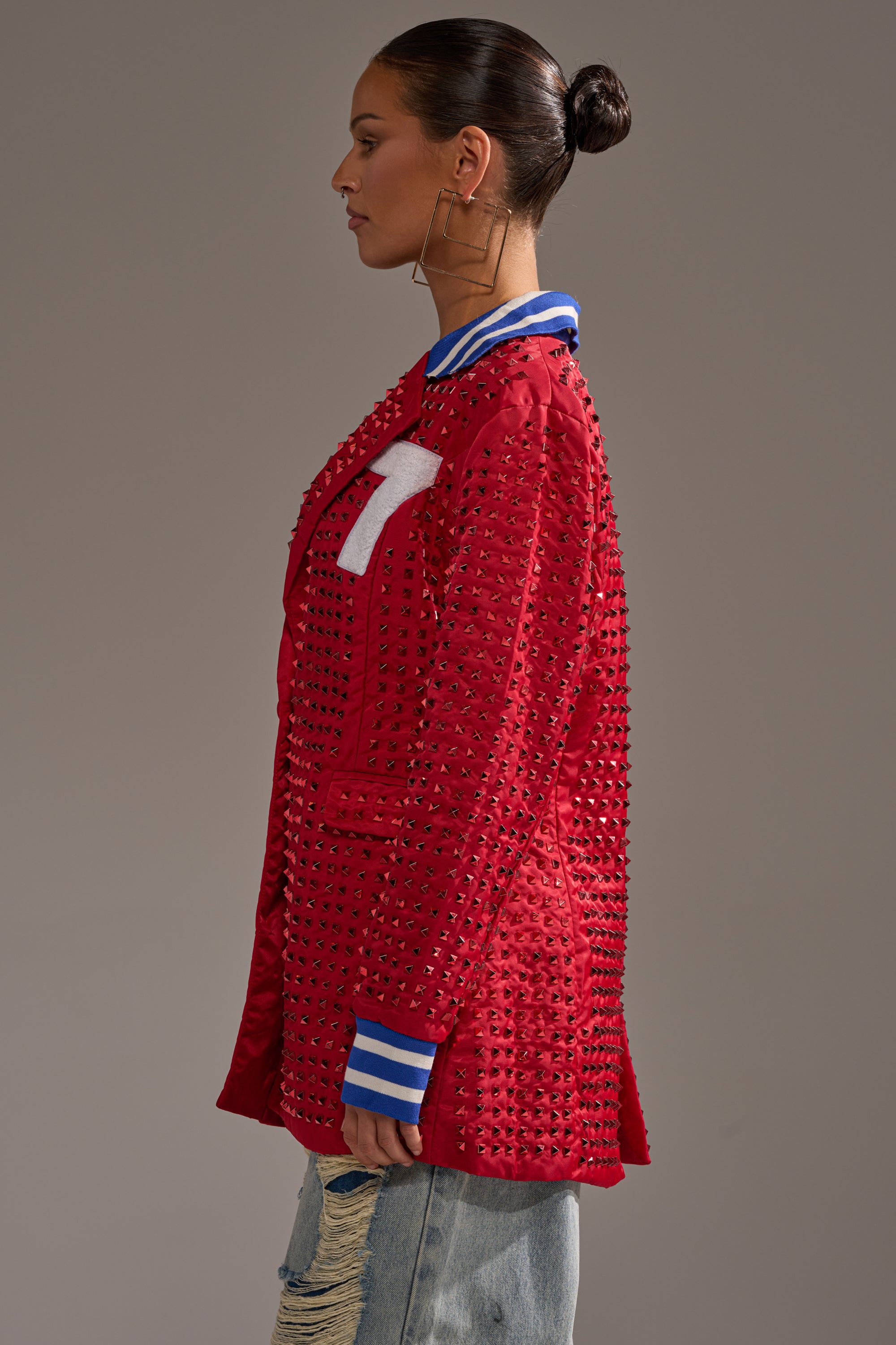 A woman in profile wears the FIRST ROUND DRAFT PICK STUDDED BLAZER IN RED with a white "T" patch, blue-and-white striped cuffs and collar, paired with ripped light blue jeans. Her hair is in a sleek bun and she sports large square hoop earrings.
