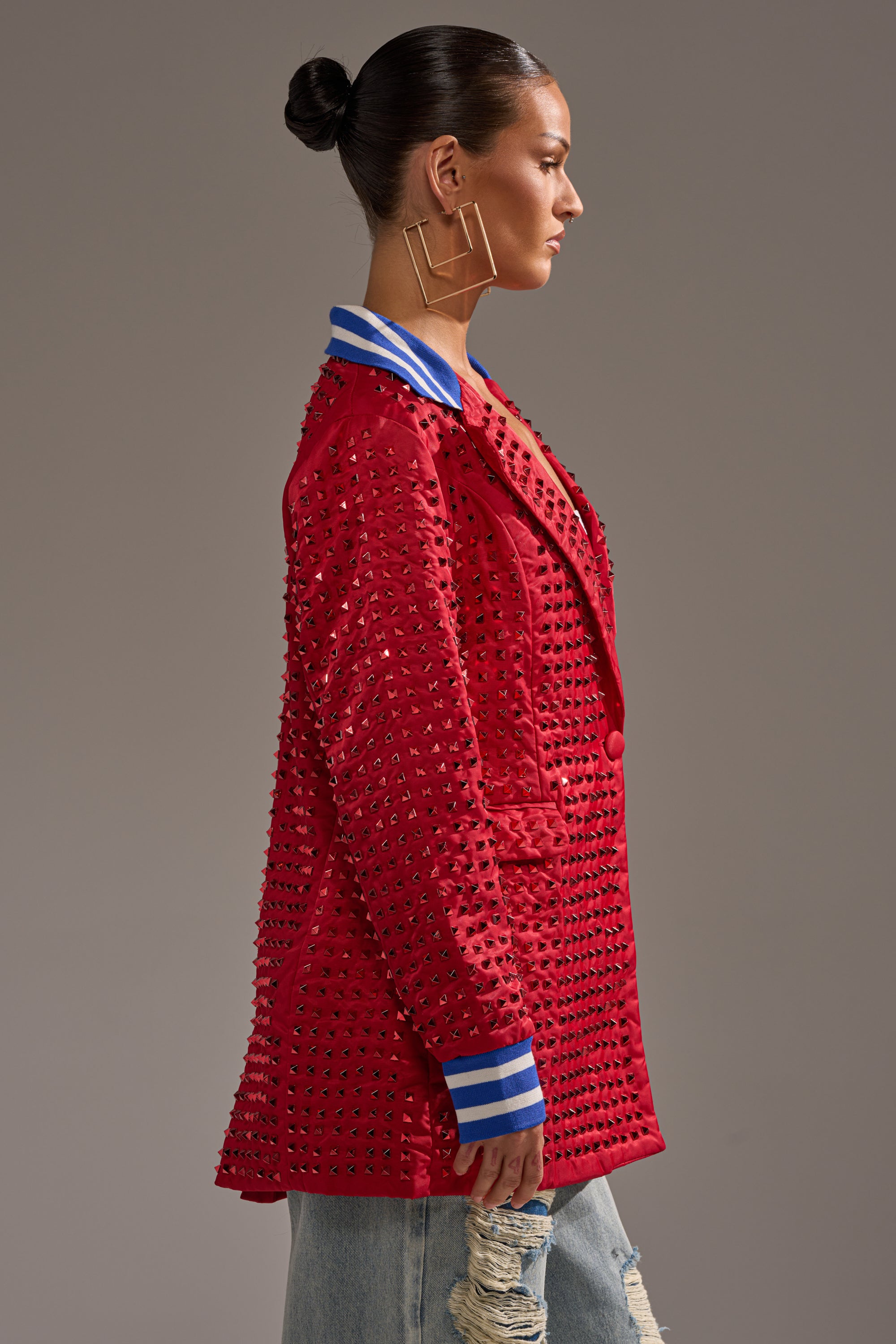 A woman with a sleek bun models the FIRST ROUND DRAFT PICK STUDDED BLAZER IN RED, featuring a blue and white striped collar and cuffs, paired with large square hoop earrings and ripped light-wash jeans, standing in profile.