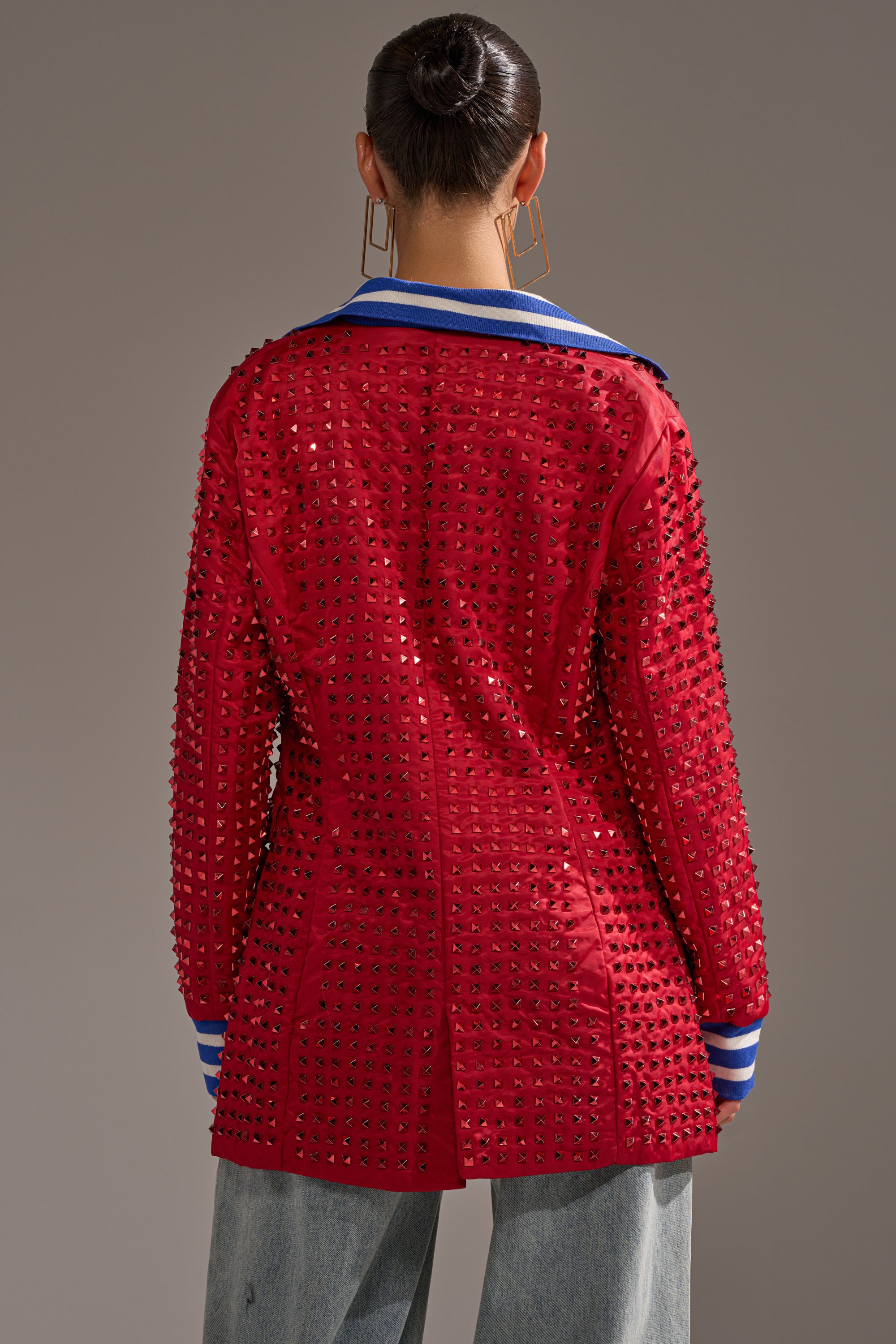 A person with dark hair in a bun, large square earrings, and light blue pants is seen from the back wearing the FIRST ROUND DRAFT PICK STUDDED BLAZER IN RED, featuring blue and white striped cuffs and collar, against a plain background.