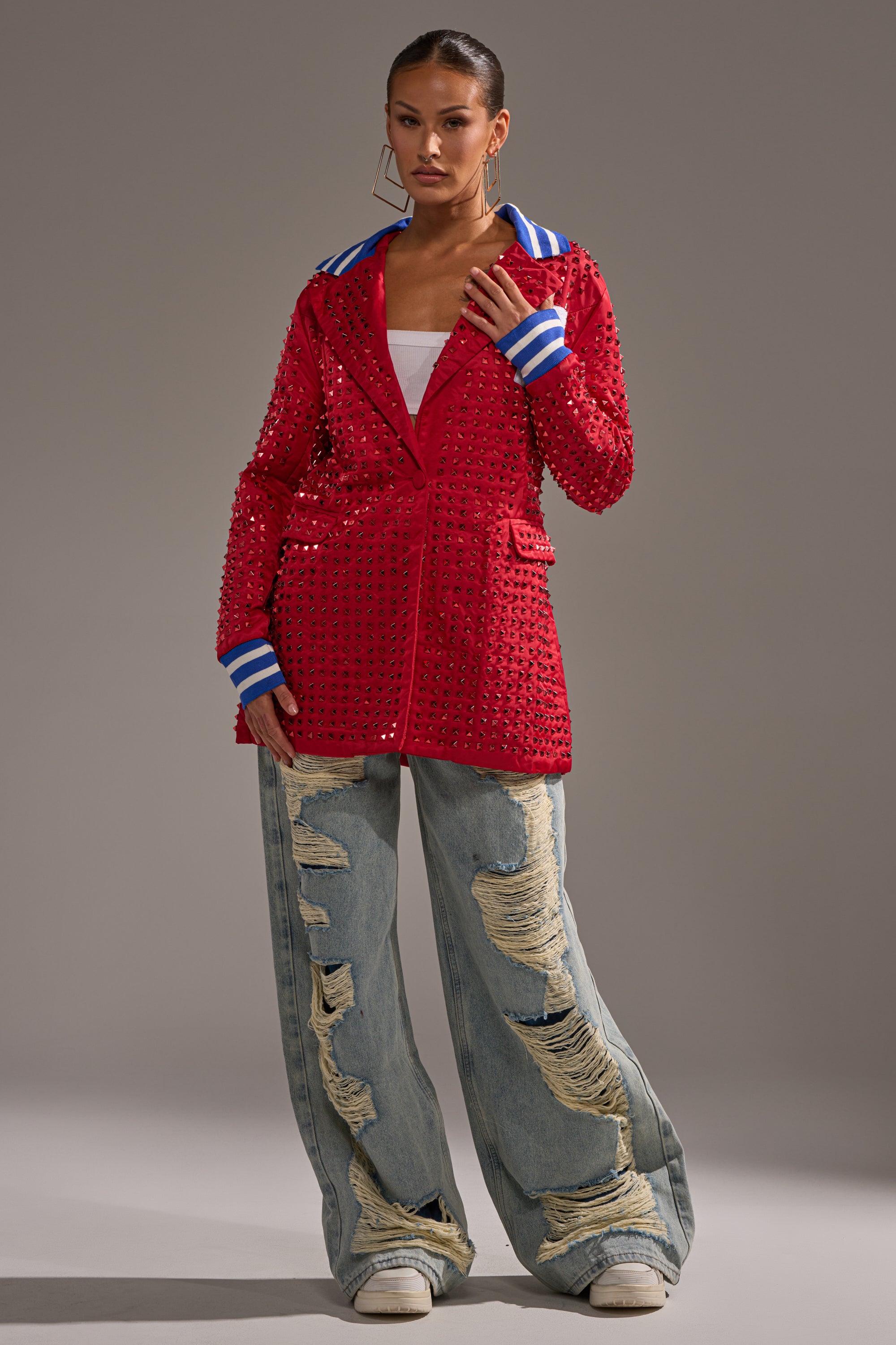 A woman wears the FIRST ROUND DRAFT PICK STUDDED BLAZER IN RED with blue and white striped cuffs, a white top, large hoop earrings, and distressed baggy jeans as she stands confidently against a plain gray background.