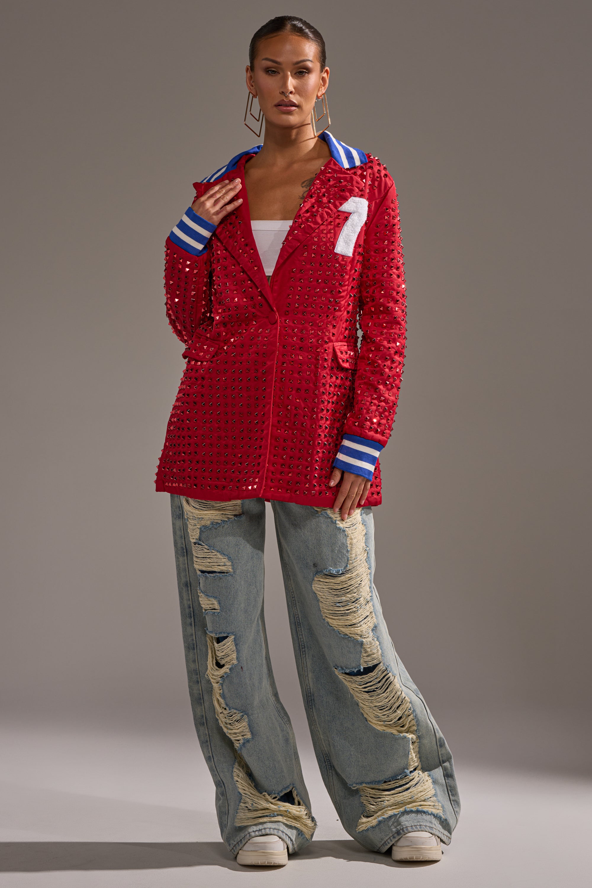 A woman faces forward against a plain background, wearing the FIRST ROUND DRAFT PICK STUDDED BLAZER IN RED with blue-and-white striped cuffs and a white number 7, paired with a white top and distressed jeans.