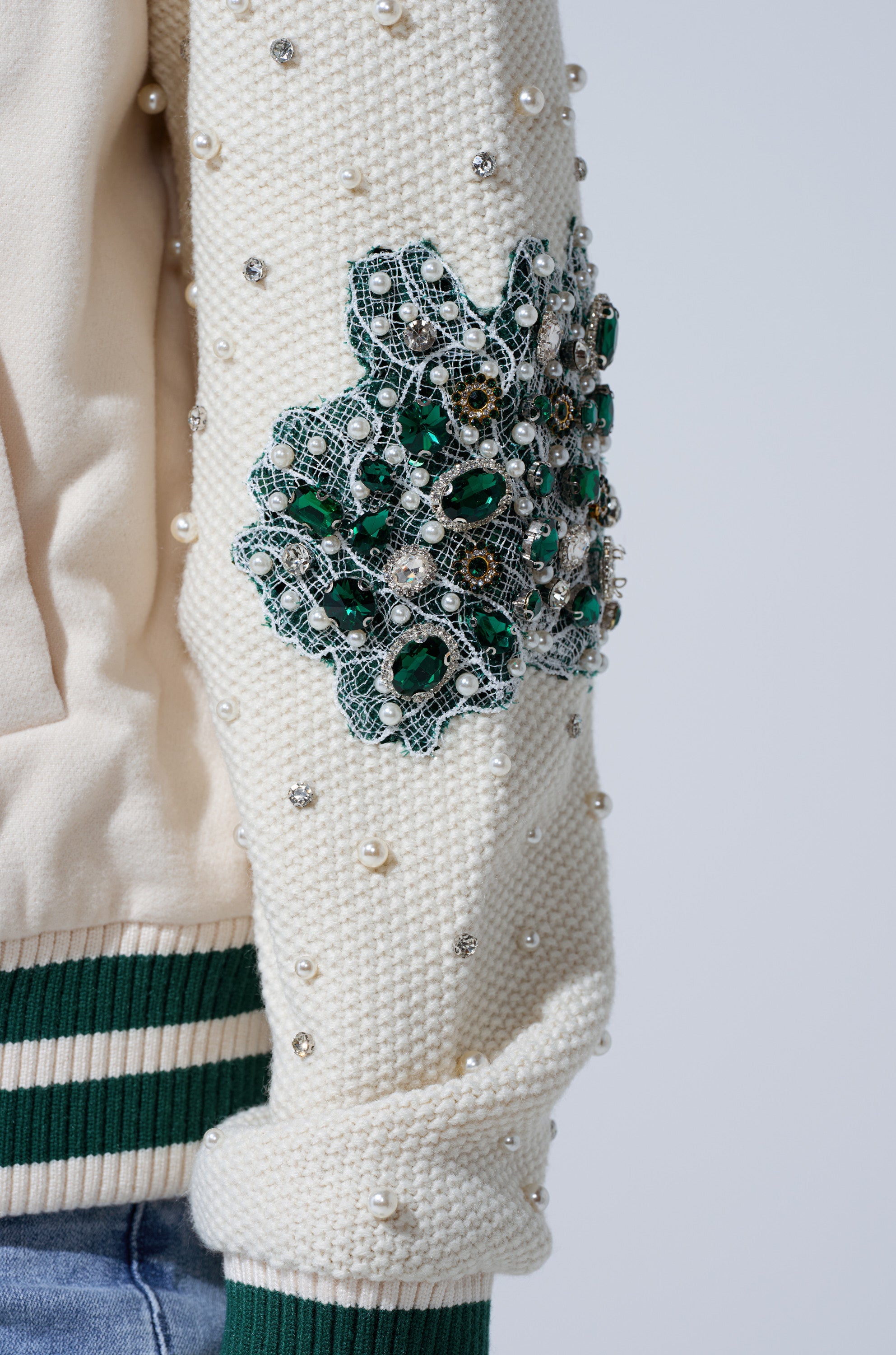 A close-up of the BETTER LUCK NEXT TIME EMBELLISHED BOMBER's cream sleeve shows pearl and silver studs, a green and silver beaded floral patch, and a cuff with green and white stripes.