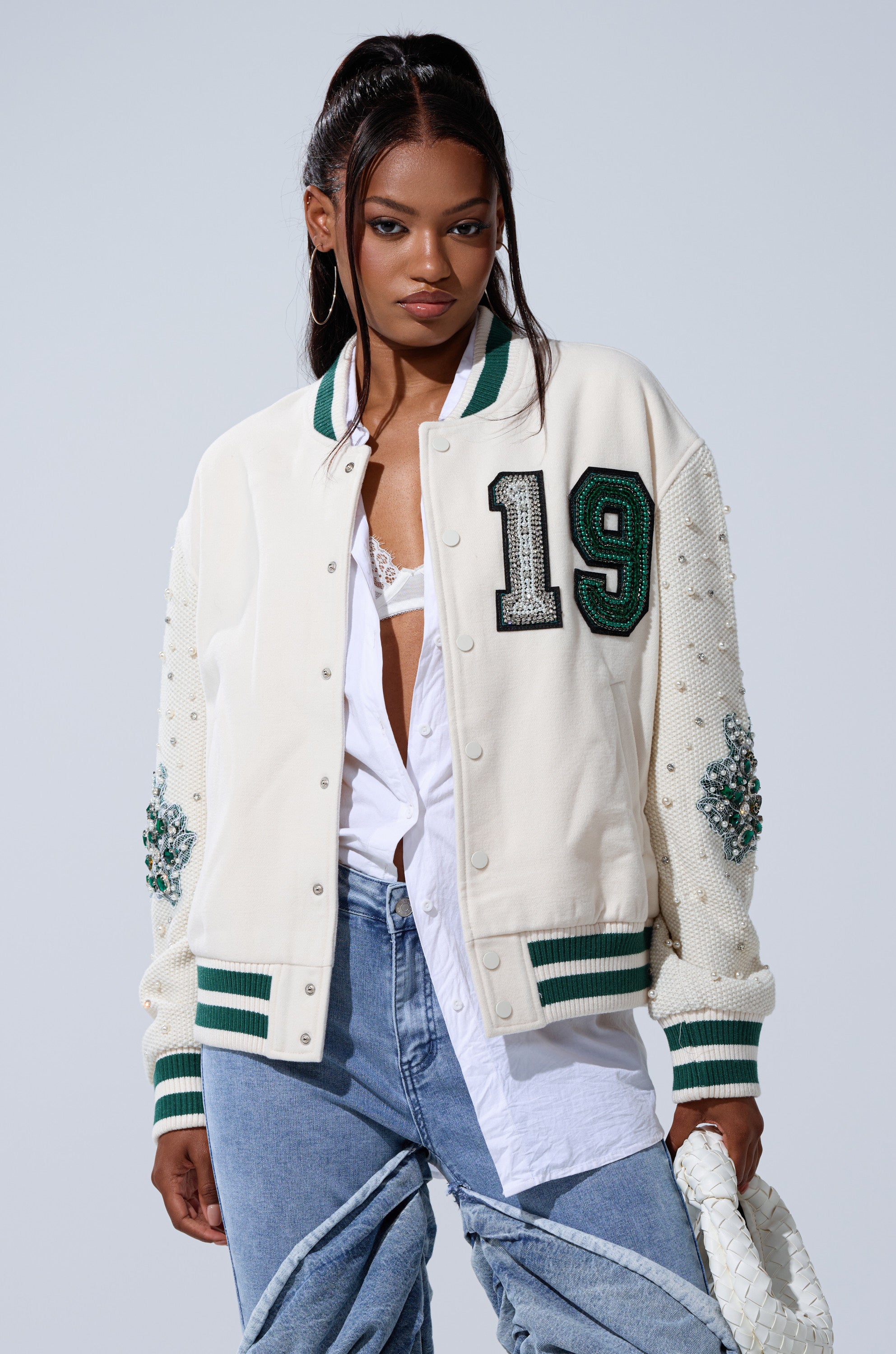 Wearing the BETTER LUCK NEXT TIME EMBELLISHED BOMBER with "19" on the chest, a white shirt, white lace bralette, light blue jeans, hoop earrings, and a woven white clutch, she poses confidently.