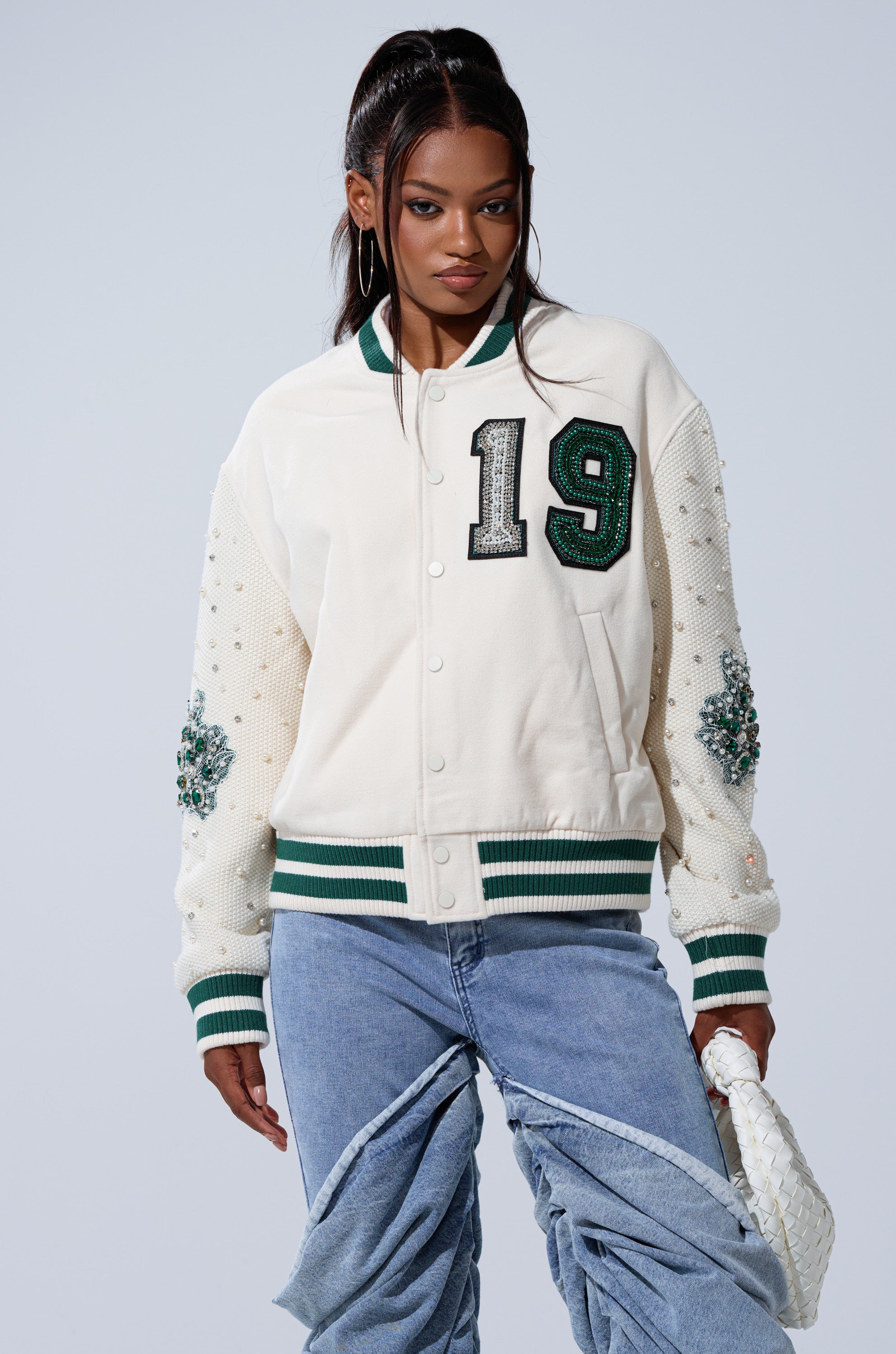 A woman with long dark hair in a high ponytail wears the BETTER LUCK NEXT TIME EMBELLISHED BOMBER, featuring pearls, rhinestones, green accents, and a bold "19" patch, paired with light blue baggy jeans and a white textured clutch.