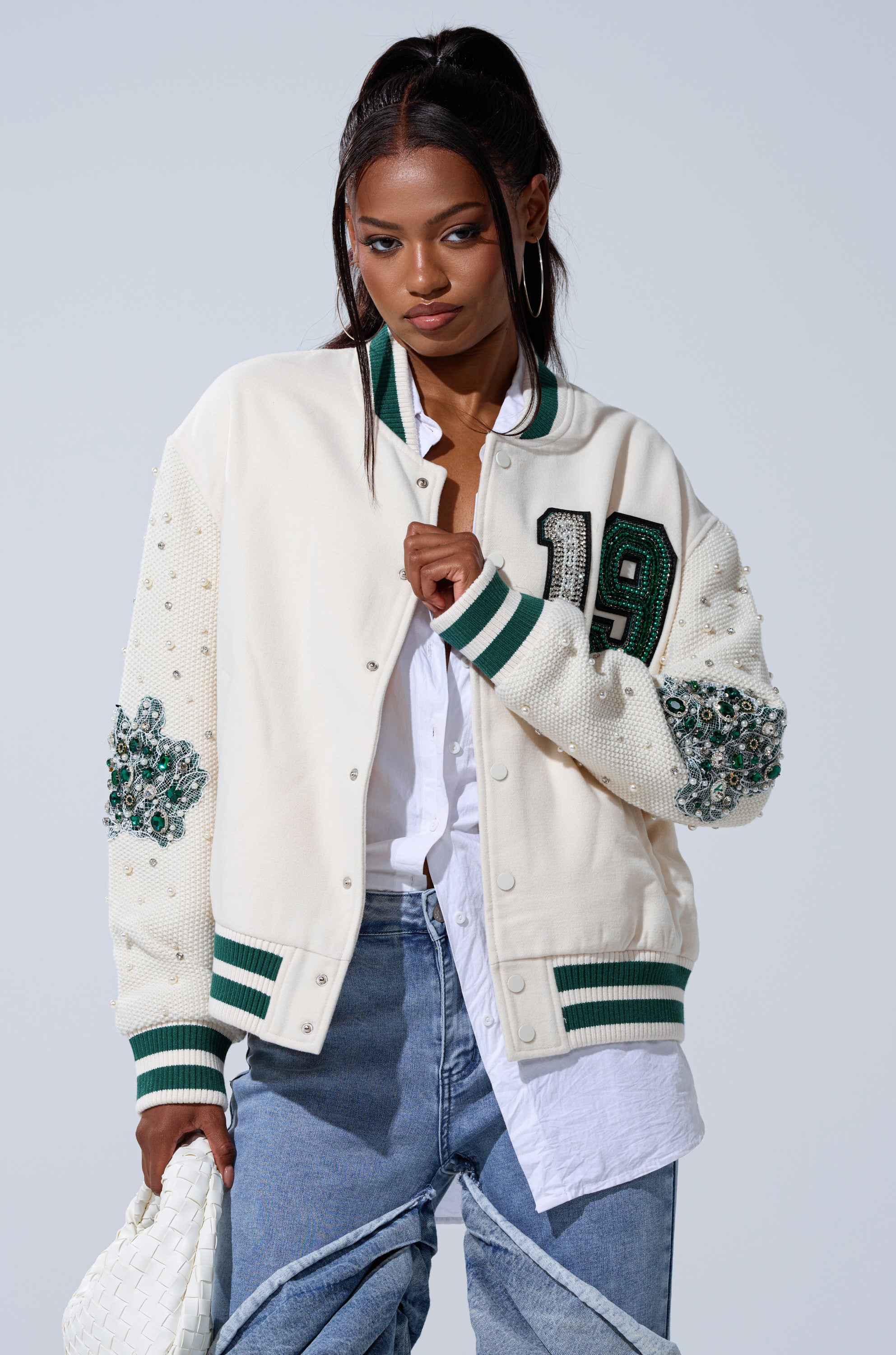 A woman wears the BETTER LUCK NEXT TIME EMBELLISHED BOMBER, a white jacket with beaded details and the number 19 on the front, paired with a white shirt, blue jeans, and a white woven bag as she looks to the side.
