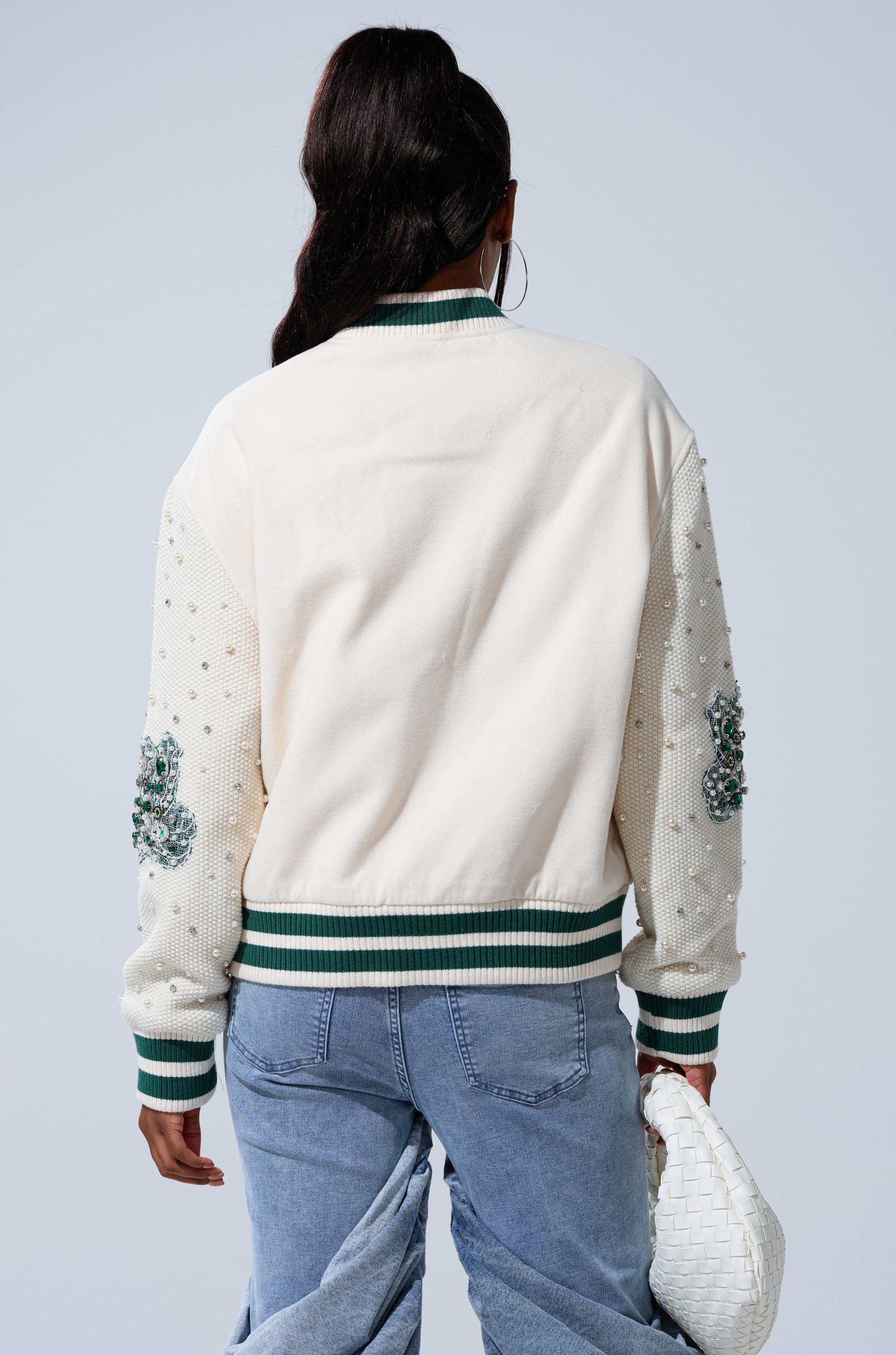 A person with long, dark hair in a ponytail is seen from behind wearing the BETTER LUCK NEXT TIME EMBELLISHED BOMBER with green and white striped trim, beaded floral sleeves, light blue jeans, and holding a white woven handbag.