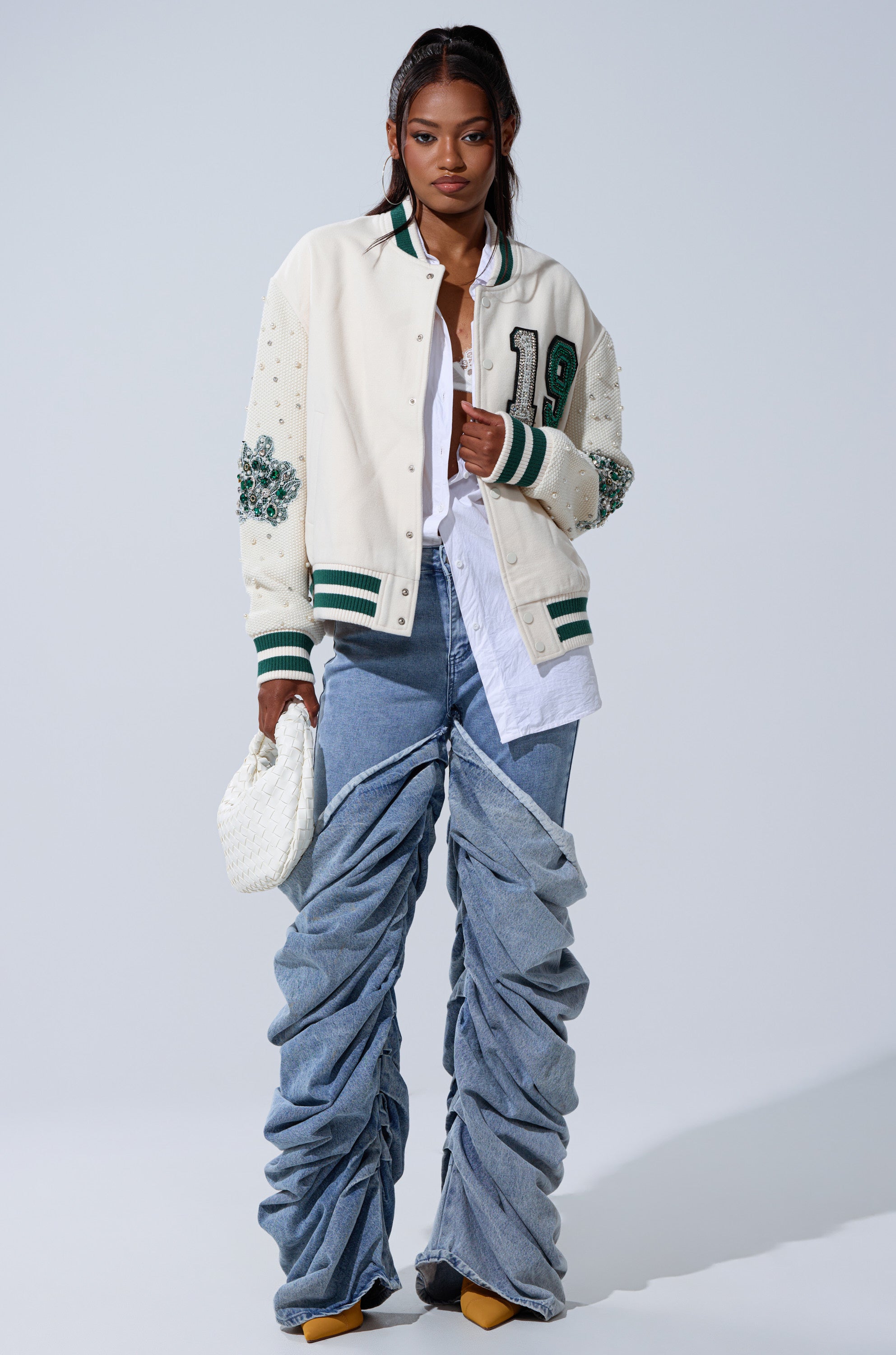 A stylish woman poses against a plain background, wearing the BETTER LUCK NEXT TIME EMBELLISHED BOMBER with green accents, a white shirt, ruched wide-leg jeans, yellow shoes, and carrying a textured white handbag.