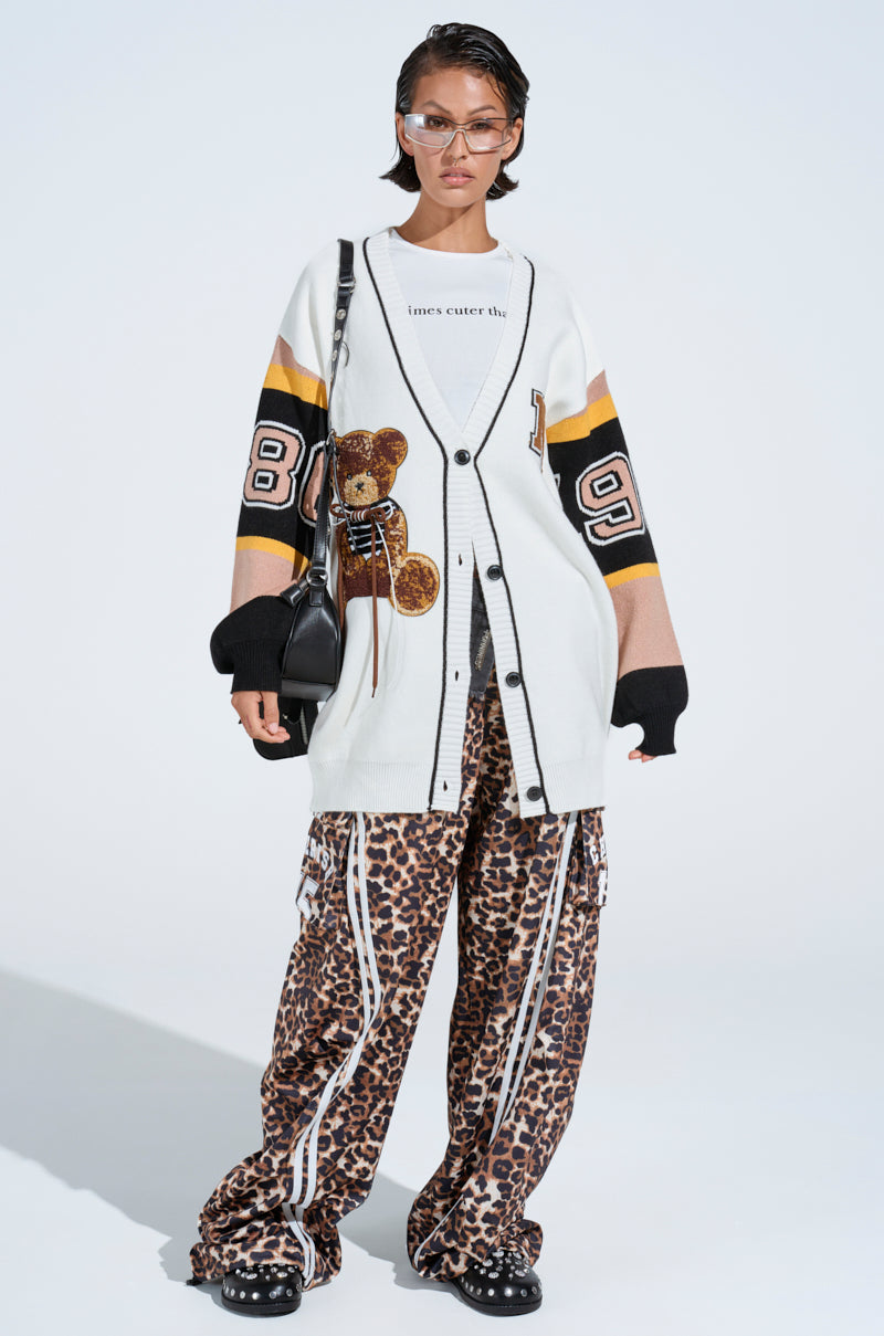 A person models a casual outfit: a long white patterned cardigan with a teddy bear patch, white graphic tee, CHERISH CHEETAH PANT—oversized high-rise with elastic waistband, and black platform sneakers. They accessorize with a black handbag and tinted sunglasses.