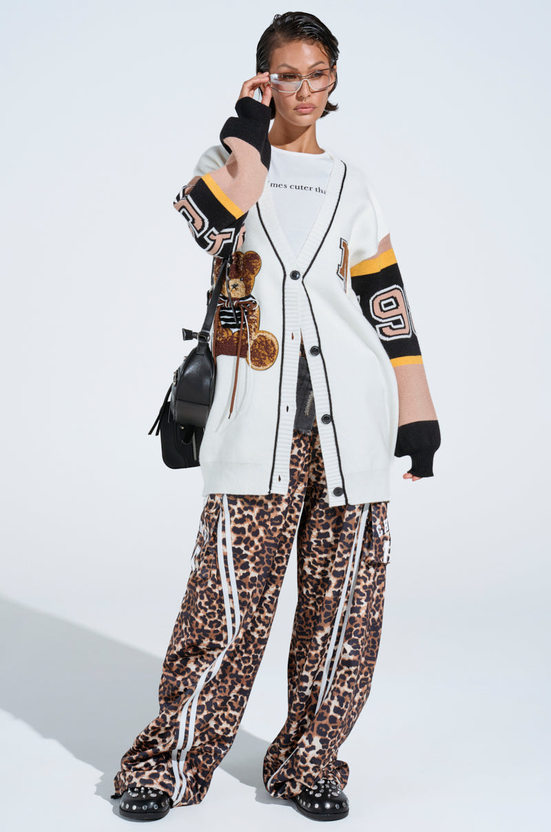 A person poses confidently in a long white cardigan with black trim and graphic designs, wearing CHERISH CHEETAH PANT featuring a high-rise fit and stretchy waistband. They adjust their sunglasses while holding a black bag over one shoulder against a plain white background.
