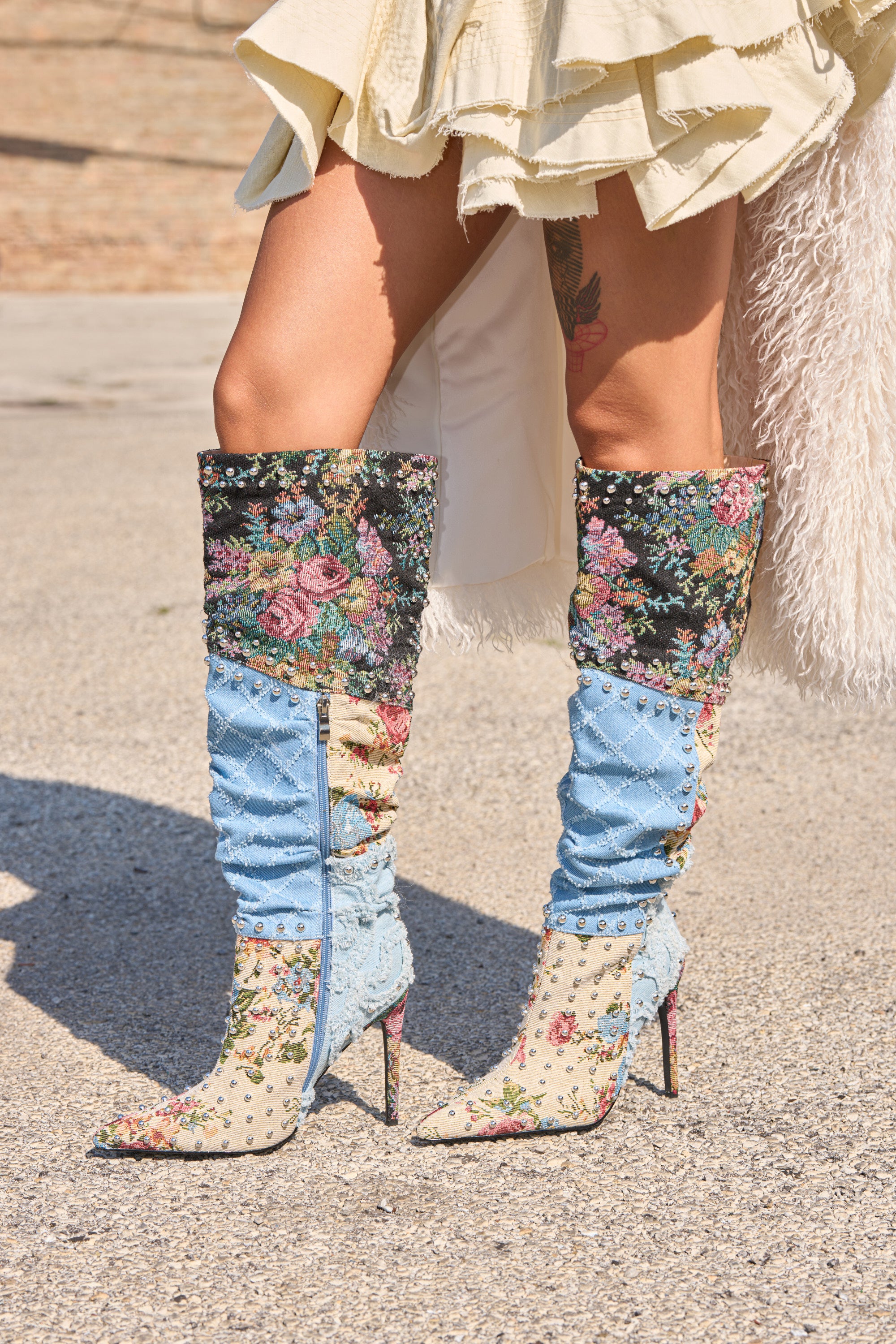 A person wearing the AZALEA WANG AILA DENIM MULTI STUDDED POINTED TOE STILETTO BOOT, styled with a cream ruffled skirt and showing a thigh tattoo, stands on a paved surface.