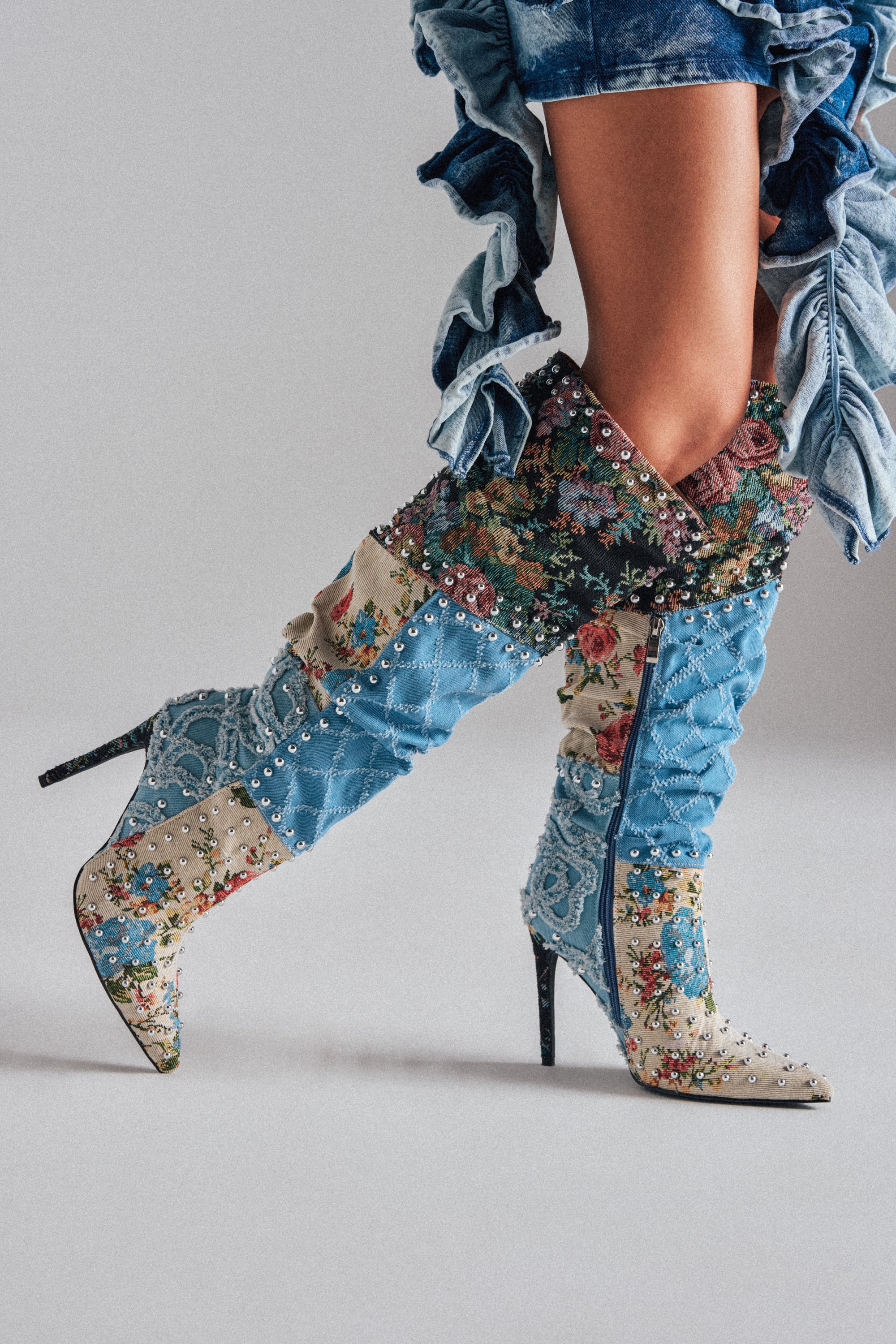 A person wearing AZALEA WANG AILA Denim Multi Studded Pointed Toe Stiletto Boots—knee-high, patchwork denim with studs and ruffles—styled with frayed denim shorts.