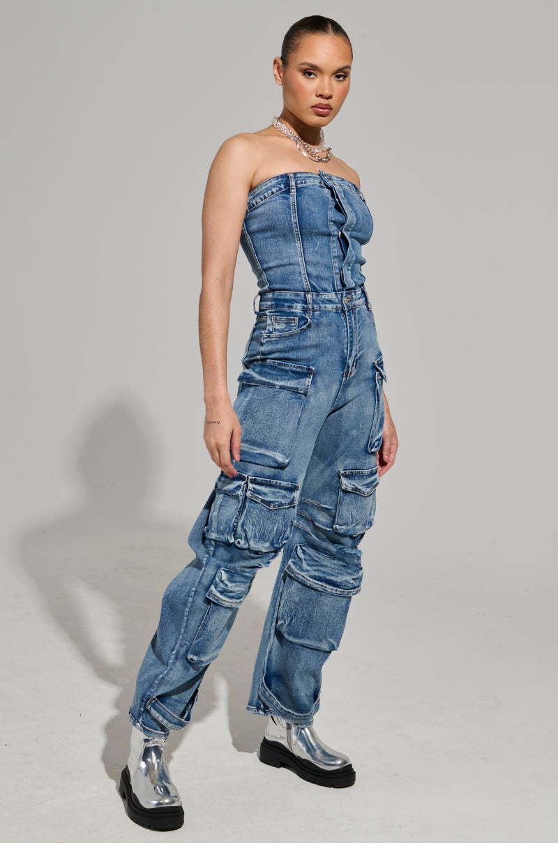 A person exudes confidence in the ROUND IT UP DENIM JUMPSUIT, showcasing cargo-style pockets and distressed detailing. With slicked-back hair, they complement the outfit flawlessly with shiny silver ankle boots against a simple background.