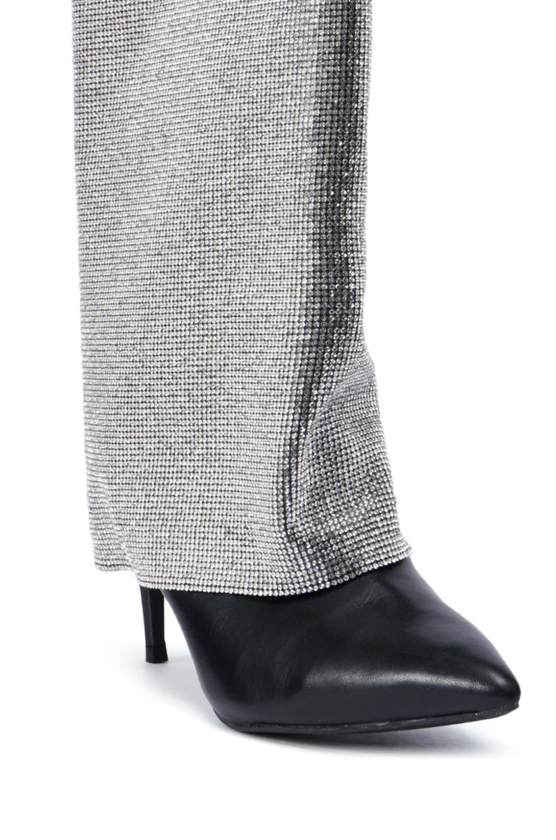 A close-up of a person wearing gray textured pants with a shimmering stripe down the side. They pair them effortlessly with the AZALEA WANG Aisling Black Sparkle Covered Heel Boot, adding a touch of elegance to the ensemble. 5