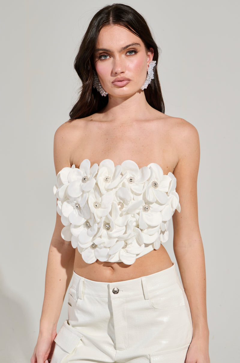 A woman with long dark hair wears a strapless FLOWER HEAVEN TOP, adorned with 3D flower appliques and styled with white pants. She completes her look with statement earrings, while the plain background accentuates the chic cropped silhouette of her fashion-forward outfit.