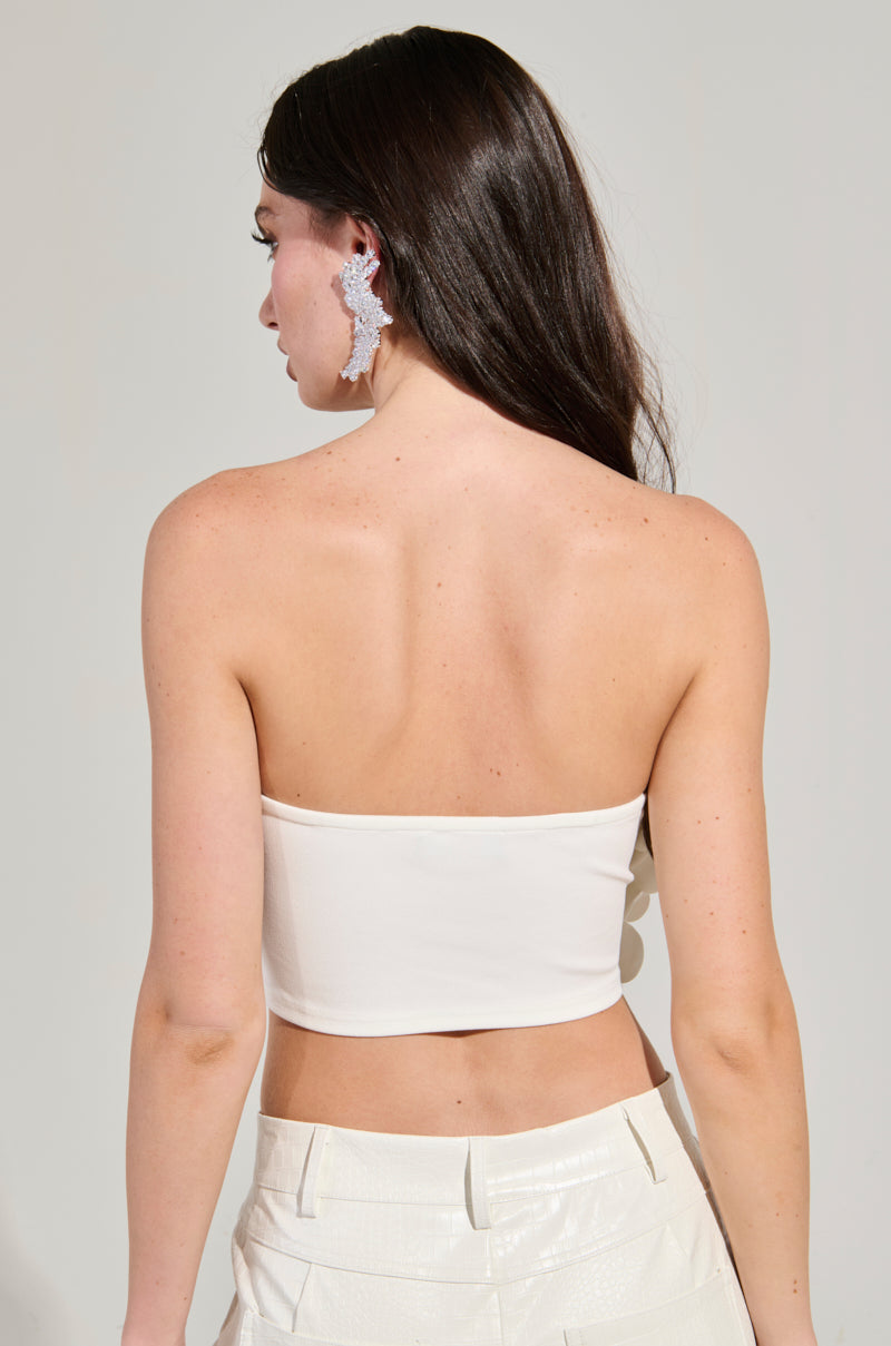 A woman with long, dark hair is facing away, dressed in the FLOWER HEAVEN TOP—a white strapless piece embellished with 3D flower appliques—and matching pants. Her ear sparkles with a large earring, accentuating her fair skin against a plain light background.
