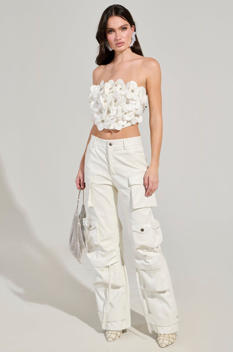 A woman stands against a simple backdrop, showcasing the FLOWER HEAVEN TOP, featuring its strapless design adorned with 3D flower appliques. She pairs this top with white cargo pants and carries a glittery silver handbag. Her ensemble is finished with large hoop earrings and patterned heels, offering a stylish cropped silhouette.