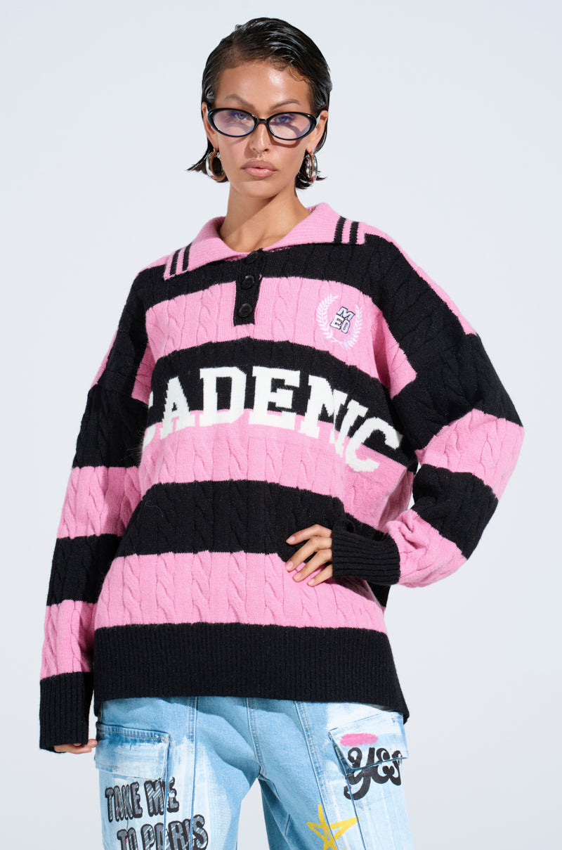 A person with short, slicked-back hair confidently poses in an oversized ACADEMIC STRIPE SWEATER with "ADENIC" on the front, black cat-eye sunglasses, and light blue jeans featuring colorful patches.