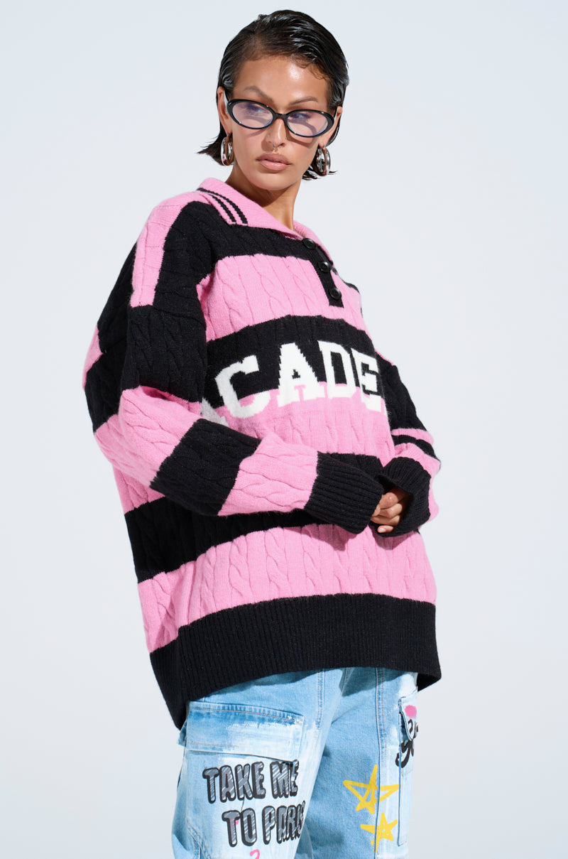Someone wears the ACADEMIC STRIPE SWEATER, featuring "ACADE" text, paired with blue jeans adorned with colorful patches and writing, plus black sunglasses, standing against a plain light background.