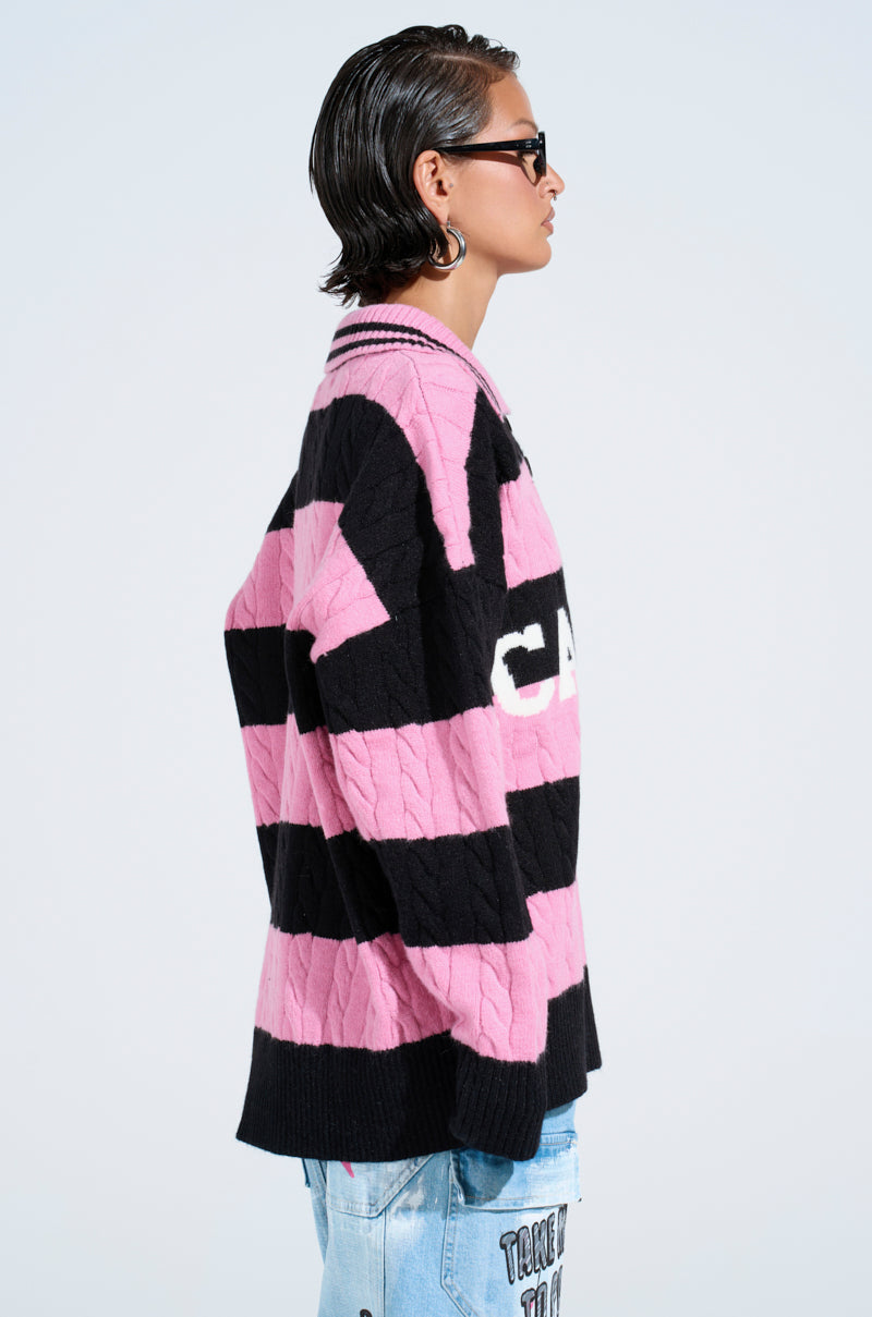A person with short, slicked-back hair and sunglasses models the oversized ACADEMIC STRIPE SWEATER in pink and black stripes, paired with patchwork light blue jeans, shown in profile against a plain light background.