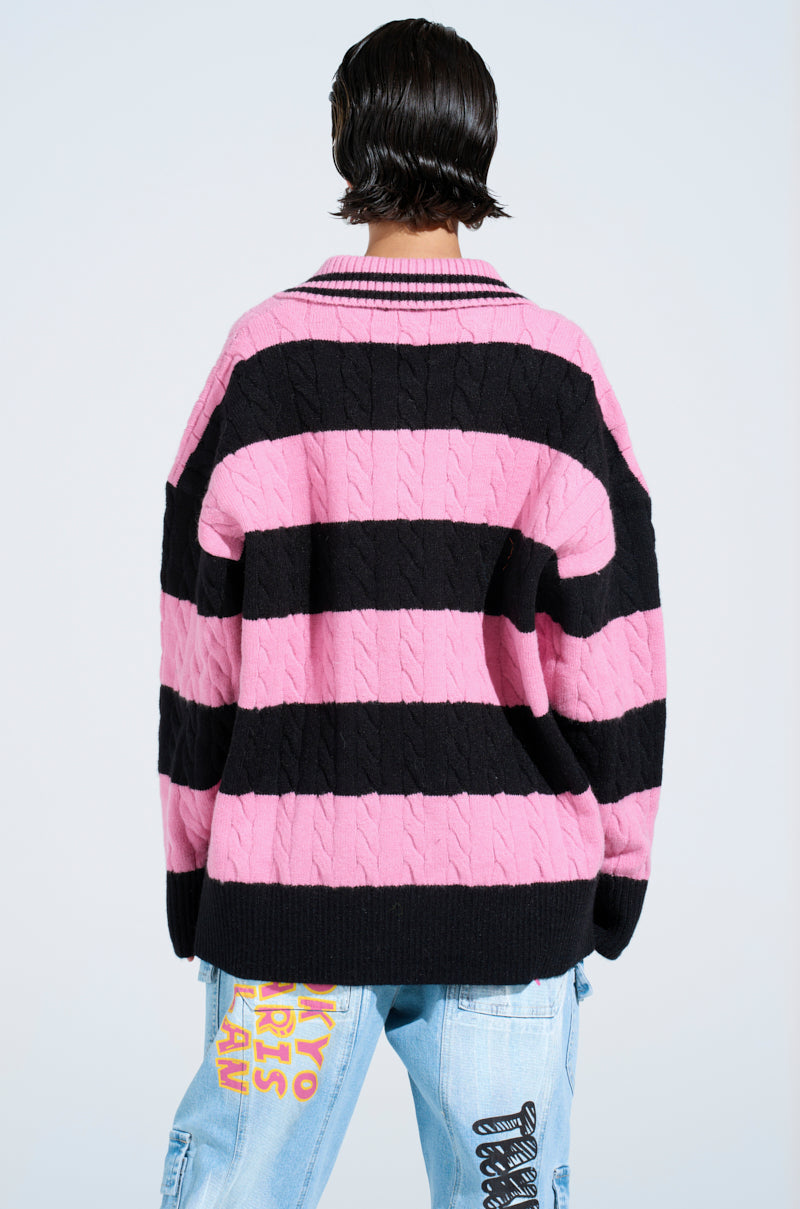 A person with short dark hair, seen from behind, wears the ACADEMIC STRIPE SWEATER in pink and black cable knit and light blue jeans featuring colorful graphics on the right leg. The background is plain and light-colored.