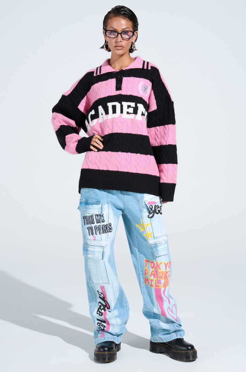 Person in an oversized pink and black ACADEMIC STRIPE SWEATER, light blue graffiti-print baggy jeans, black chunky shoes, and glasses stands against a plain light background.