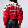 A person with long brown hair is seen from the back, wearing a red and white LIVING FREE BOMBER jacket with "Riding High" and "Rodeo" printed on it. The person is also wearing black leather pants.