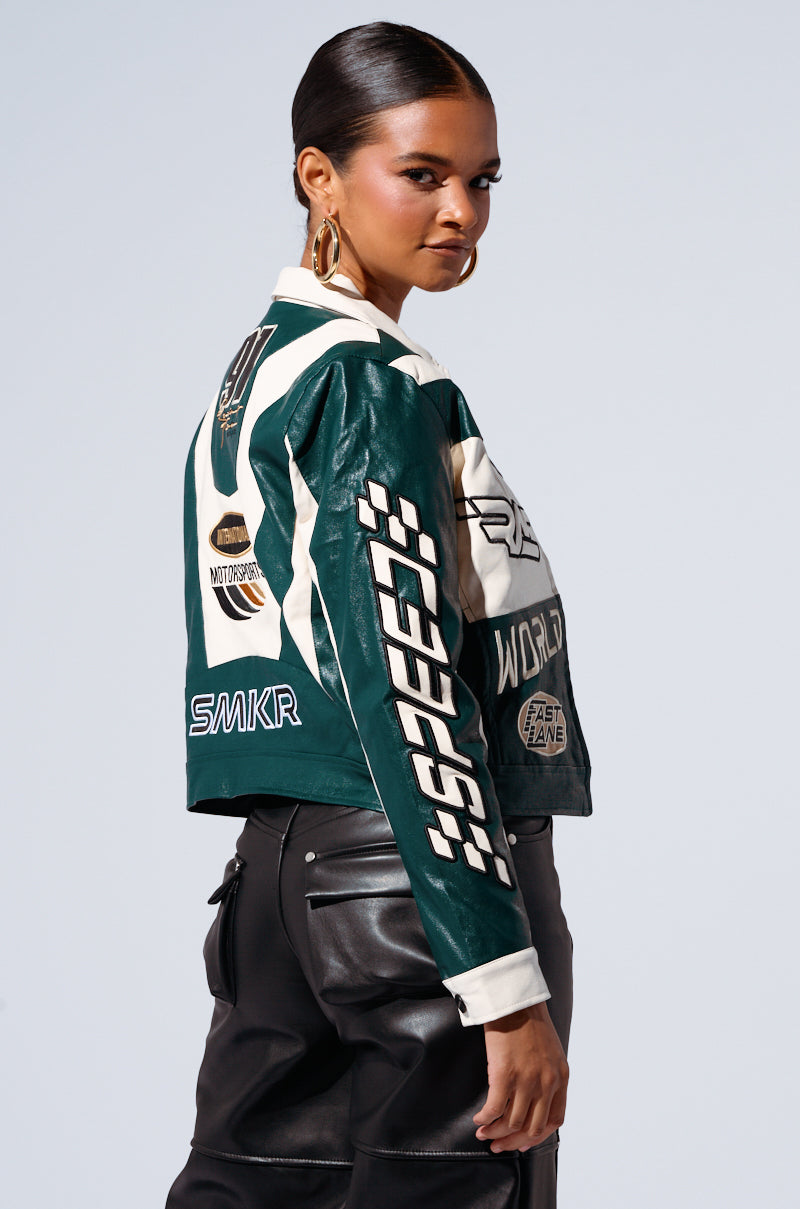A person with slicked-back hair stands facing slightly to the side, wearing the HATE THE PLAYER BOMBER, a stylish green and white motorcycle-style jacket with colorblock paneling and various patches. Paired with black high-waisted faux leather pants, they look over their shoulder with a confident expression.