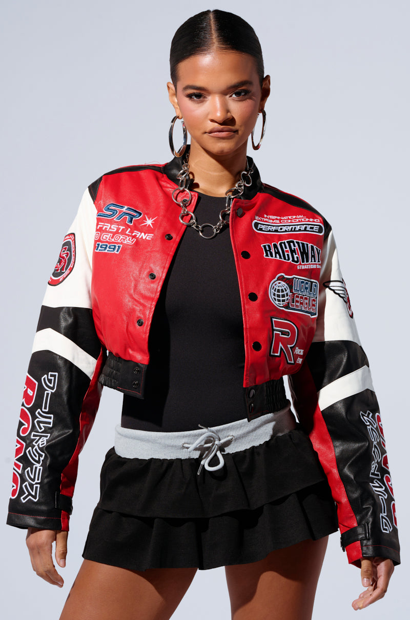 A woman is dressed in the MOTOR SPORT RACER BOMBER, a stylish jacket adorned with embroidered patches in vibrant shades of red, black, and white. She complements this look with a sleek black ensemble featuring a ruffled mini skirt and a light grey belt. Her hair is elegantly pulled back, she wears large hoop earrings, and stands against a plain background. 0