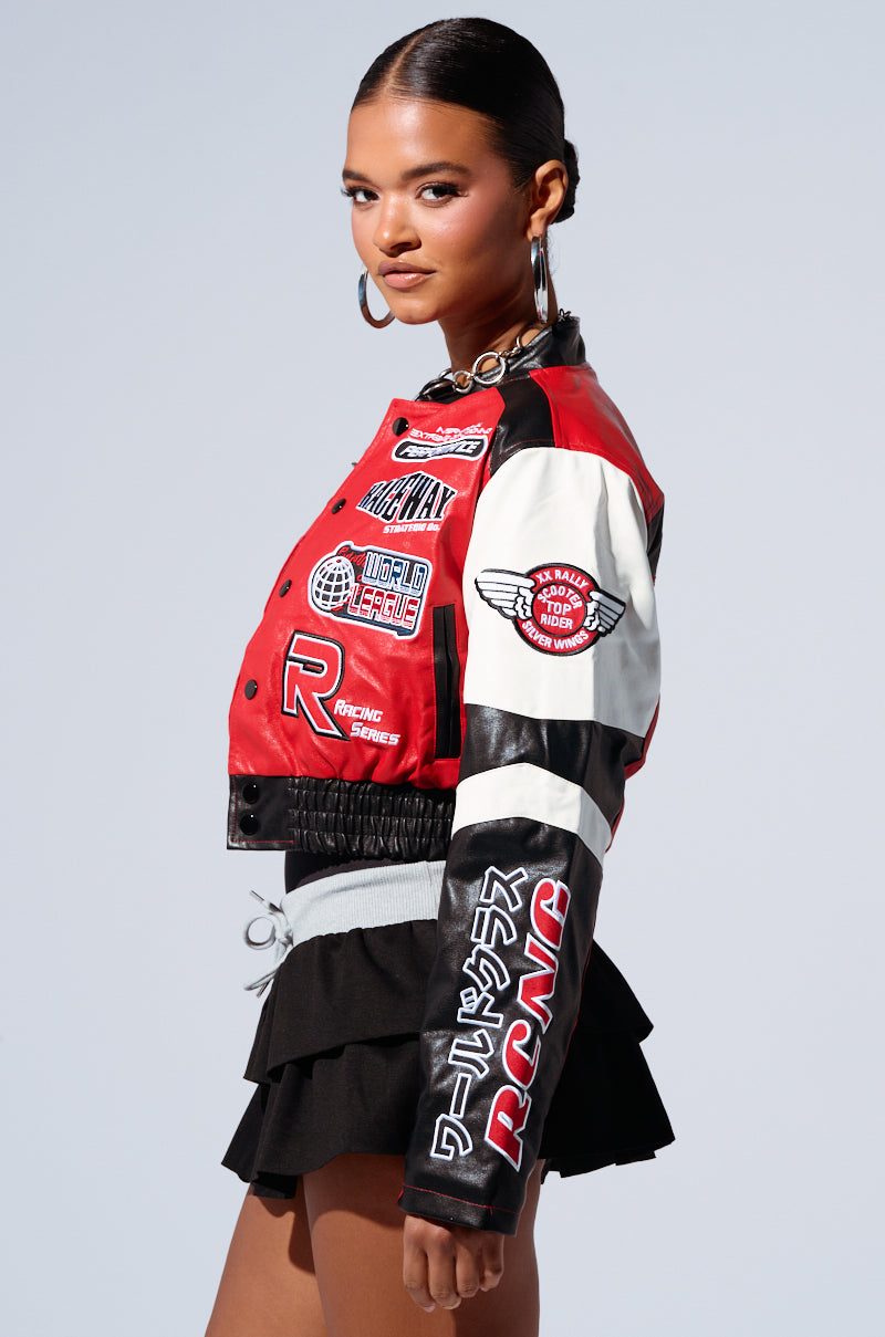 A woman poses confidently against a light grey background, wearing the MOTOR SPORT RACER BOMBER jacket adorned with various racer-inspired embroidered patches and logos such as "World Champs" and "Racing Speed." She accessorizes with hoop earrings and a black, layered mini skirt. The faux leather base of the jacket adds an edgy touch as she looks over her shoulder. 1