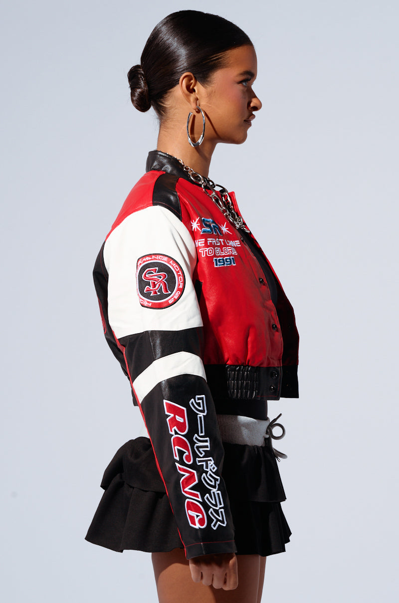 A young woman stands in a side profile against a plain light background. She is wearing a black skirt and the MOTOR SPORT RACER BOMBER jacket, which features racer-inspired embroidered patches on its faux leather base in red, black, and white varsity style. Her hair is slicked back into a neat bun, and she is adorned with large hoop earrings. 2