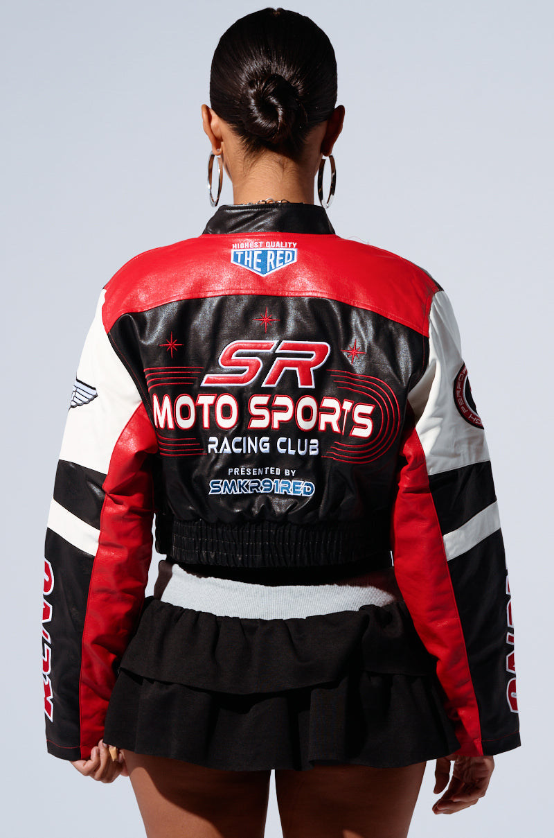 A person facing away from the camera is seen wearing the MOTOR SPORT RACER BOMBER jacket, characterized by its black, red, and white color scheme and "SR Moto Sports Racing Club" text. This faux leather jacket is adorned with various embroidered patches and logos. They have their hair tied back and are also dressed in a black skirt with ruffled layers. 3