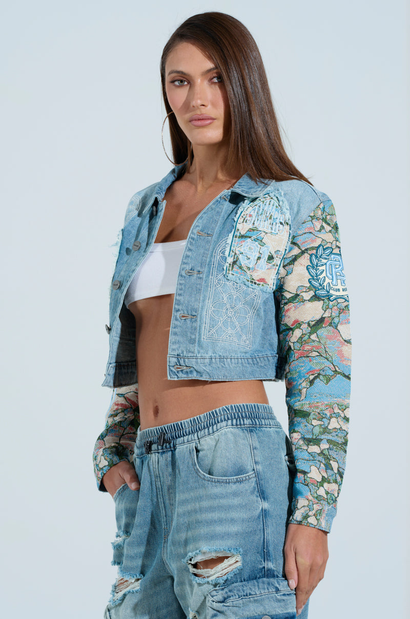 Someone flaunts the OUT OF THIS WORLD DENIM JACKET featuring graphic patches on the sleeves over a white crop top, paired with high-waisted jeans and hands casually tucked in pockets, all against a plain white background.