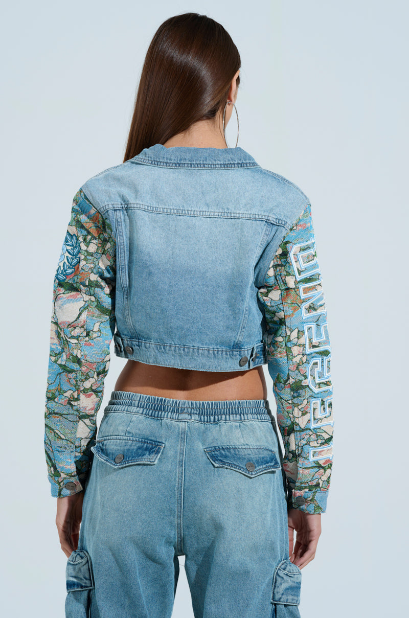 A person with long brown hair, wearing the OUT OF THIS WORLD DENIM JACKET featuring mosaic-patterned sleeves and "LEGEND" patches, pairs it with high-waisted denim cargo pants while standing against a light blue background, facing away.