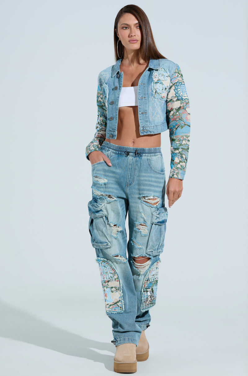 A woman wears the "Out of This World" floral patchwork denim jacket with graphic patches, a white bandeau top, and distressed denim cargo pants. Tan platform shoes complete her look as her long brown hair cascades against a light blue backdrop.
