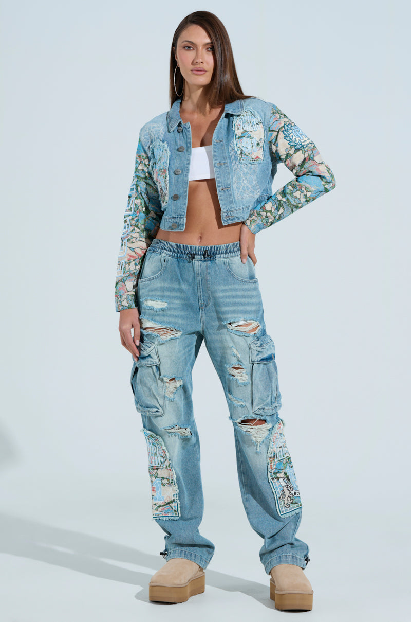 A person confidently wears the OUT OF THIS WORLD DENIM JACKET adorned with graphic patches, paired with ripped, slightly baggy jeans. Light brown platform shoes and a white cropped silhouette top complete their edgy look.
