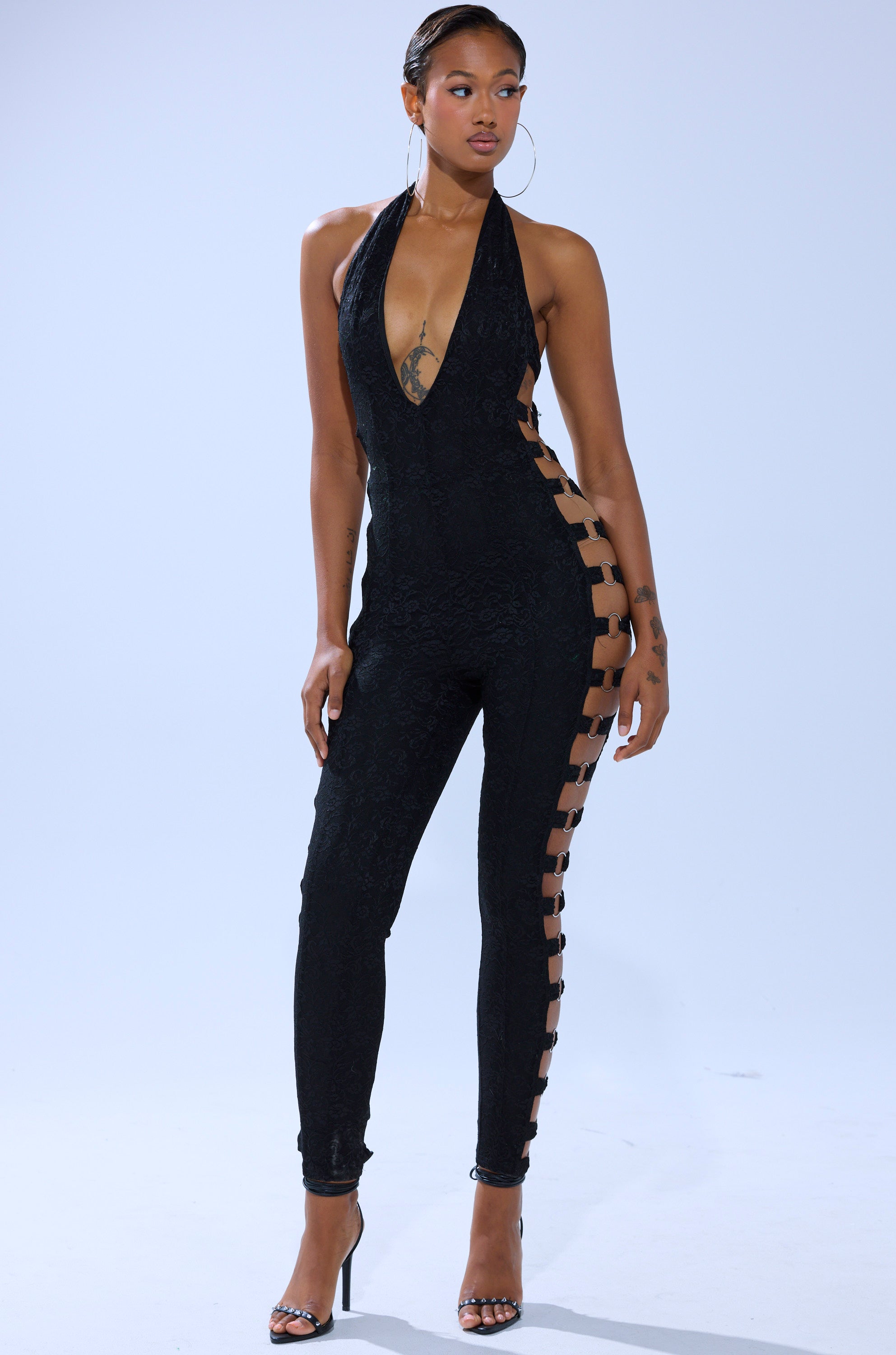 A woman models the NOT FOR EVERYONE CUTOUT LACE JUMPSUIT, showcasing its black lace design, plunging neckline, cutout sides with crisscross straps. She accessorizes with hoop earrings and high heels, her hair pulled back. 0