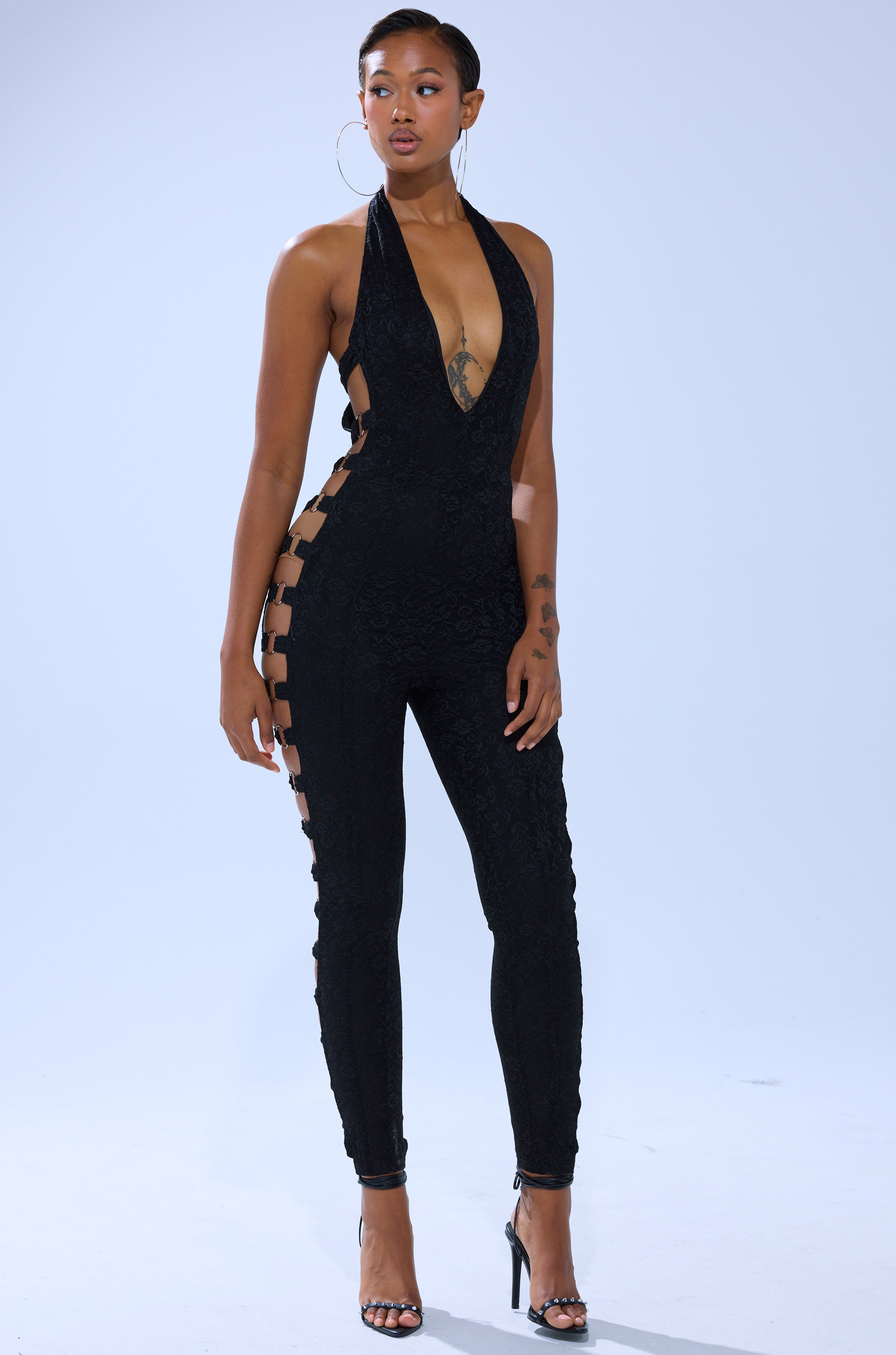 A woman stands confidently in high heels against a plain light background, wearing the NOT FOR EVERYONE CUTOUT LACE JUMPSUIT—a black, form-fitting piece featuring a deep V-neckline and bold side cutouts. 2