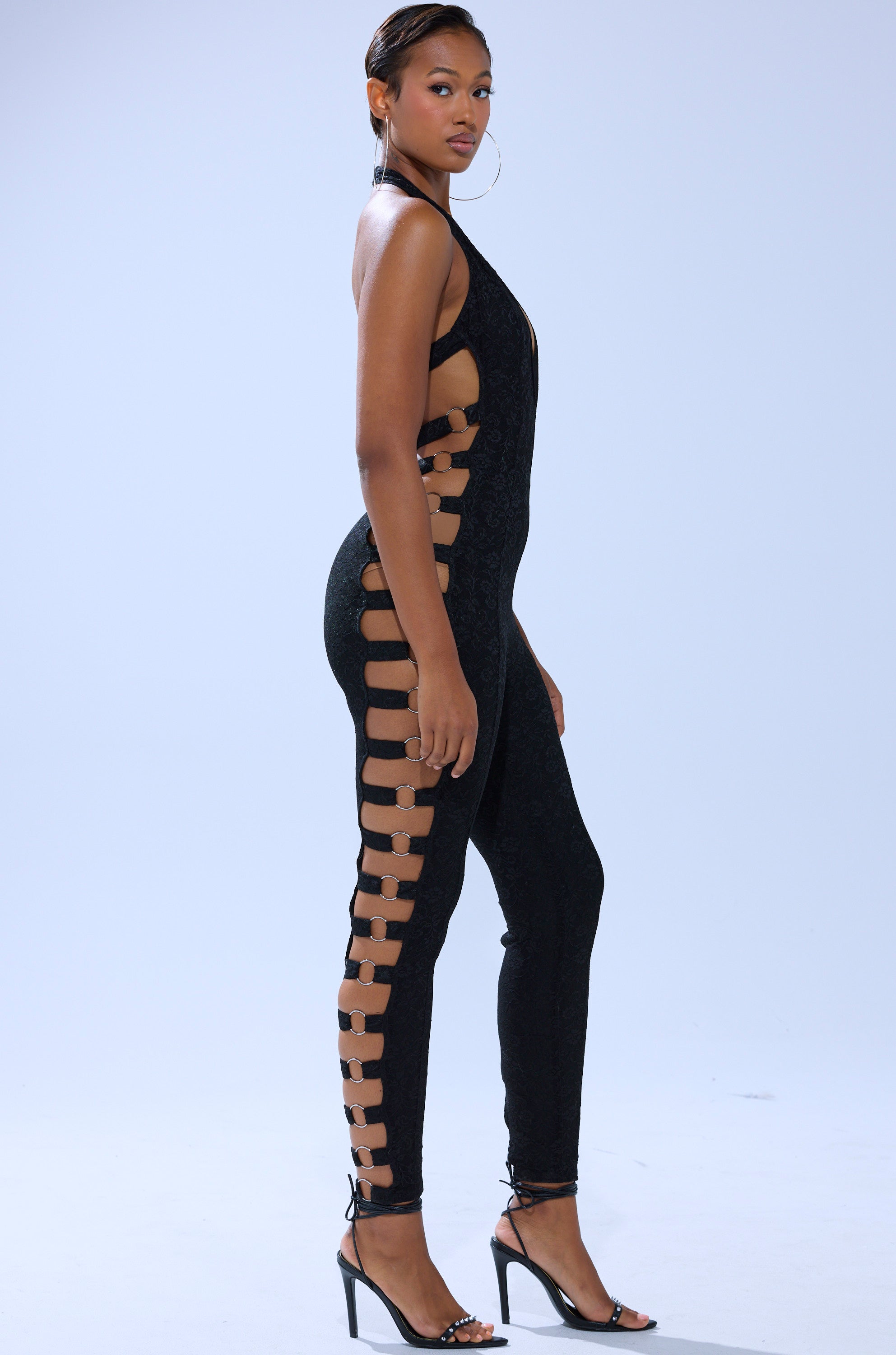 A woman models the NOT FOR EVERYONE CUTOUT LACE JUMPSUIT with side cutouts and strappy details, paired with high-heeled sandals, against a plain light background. 3