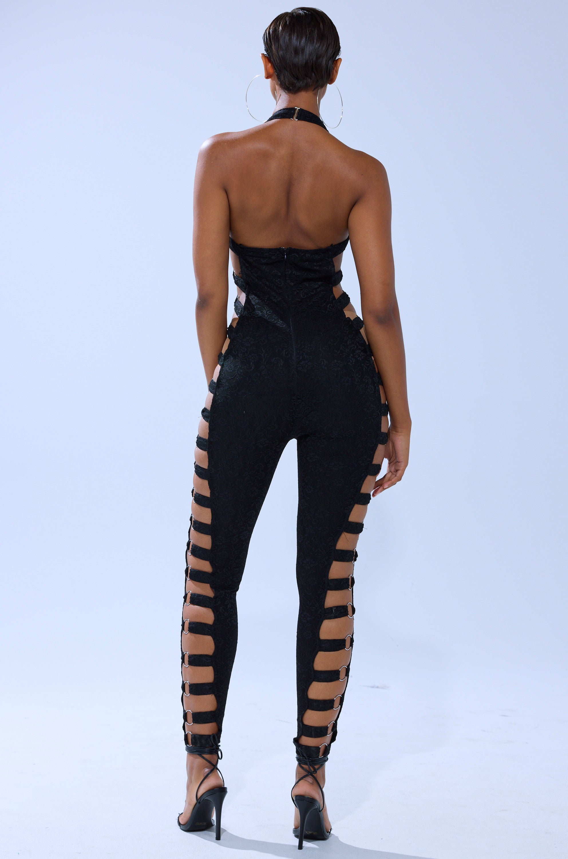 A person faces away, wearing the NOT FOR EVERYONE CUTOUT LACE JUMPSUIT featuring open sides and leg cutouts, paired with black high-heeled sandals. The backdrop is plain and light-colored. 4