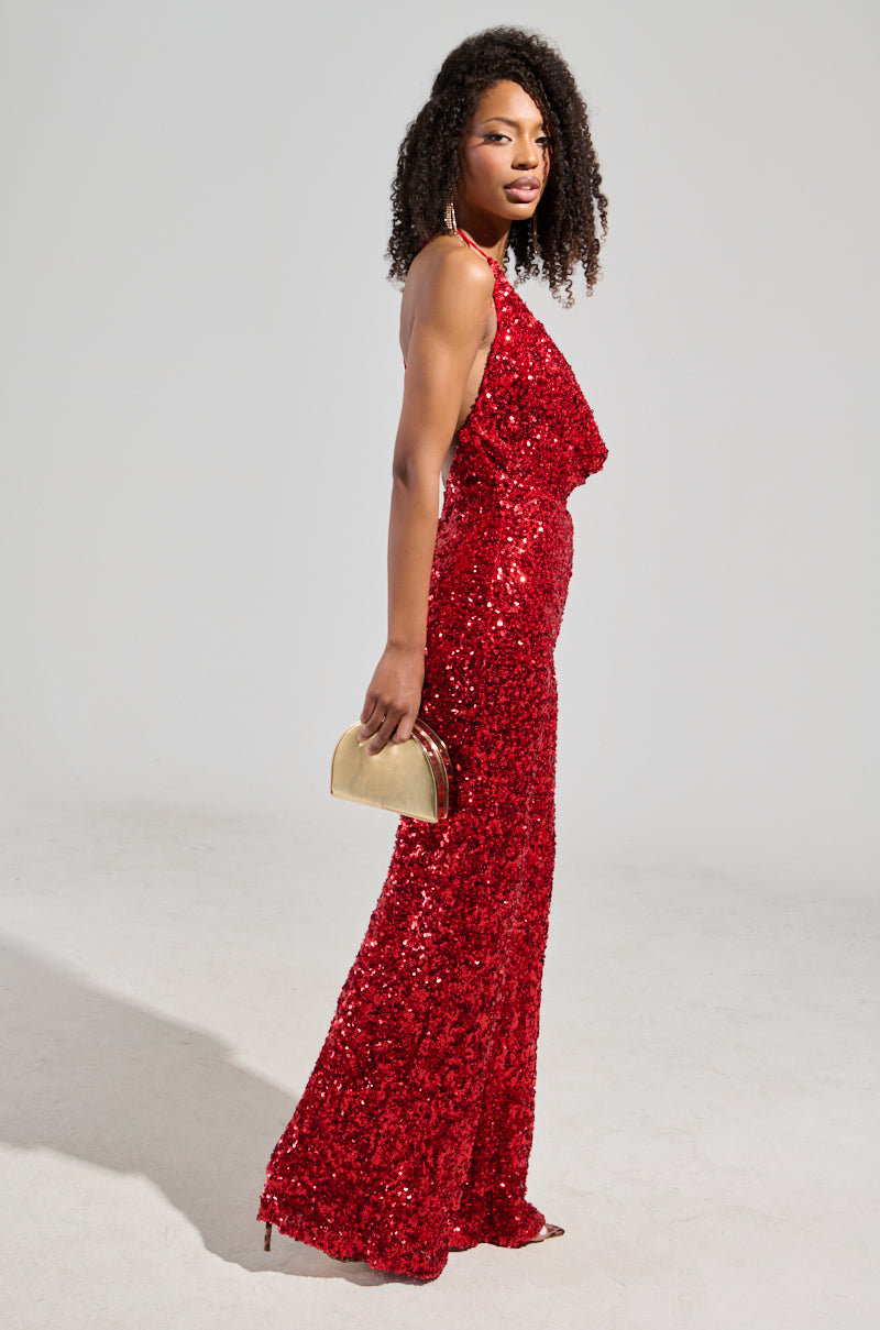 A person wearing the Jodie Scoop Neck Sequin Jumpsuit in red, with a plunging cowl neckline, holds a small gold clutch bag. Their curly hair cascades gracefully as they stand against a plain, light-colored background.