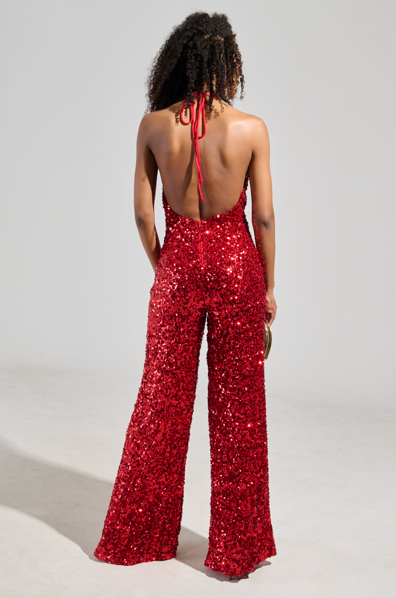 A person with curly hair stands facing away, donning the captivating JODIE SCOOP NECK SEQUIN JUMPSUIT IN RED. Its open back and halter neck design create a stunning silhouette against the backdrop of a plain, light-colored room, enhancing the ensemble's dazzling allure.