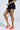 The person is wearing a black denim skirt with side pockets and the AZALEA WANG AJIT ORANGE SANDAL, noted for its novelty ball-shaped heels and satin textile upper, standing confidently with one leg slightly forward against a light gray background.