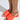 Someone wearing the AZALEA WANG AJIT ORANGE SANDAL sports bright orange open-toe mules with a satin textile upper, wide straps featuring a small logo, and a round novelty heel. The light casts a soft shadow on the floor, accentuating their vibrant color and design.
