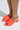 Someone wearing the AZALEA WANG AJIT ORANGE SANDAL sports bright orange open-toe mules with a satin textile upper, wide straps featuring a small logo, and a round novelty heel. The light casts a soft shadow on the floor, accentuating their vibrant color and design.