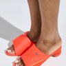 Someone wearing the AZALEA WANG AJIT ORANGE SANDAL sports bright orange open-toe mules with a satin textile upper, wide straps featuring a small logo, and a round novelty heel. The light casts a soft shadow on the floor, accentuating their vibrant color and design.