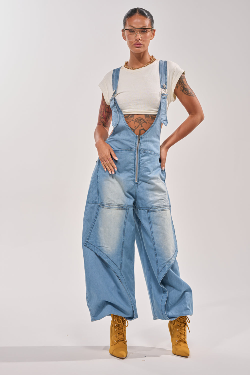 A woman confidently wears the OUT TO PLAY DENIM OVERALL JUMPSUIT over a white crop top, showing off her tattooed arms. She pairs clear protective glasses with yellow lace-up boots, standing against a plain light background. 0